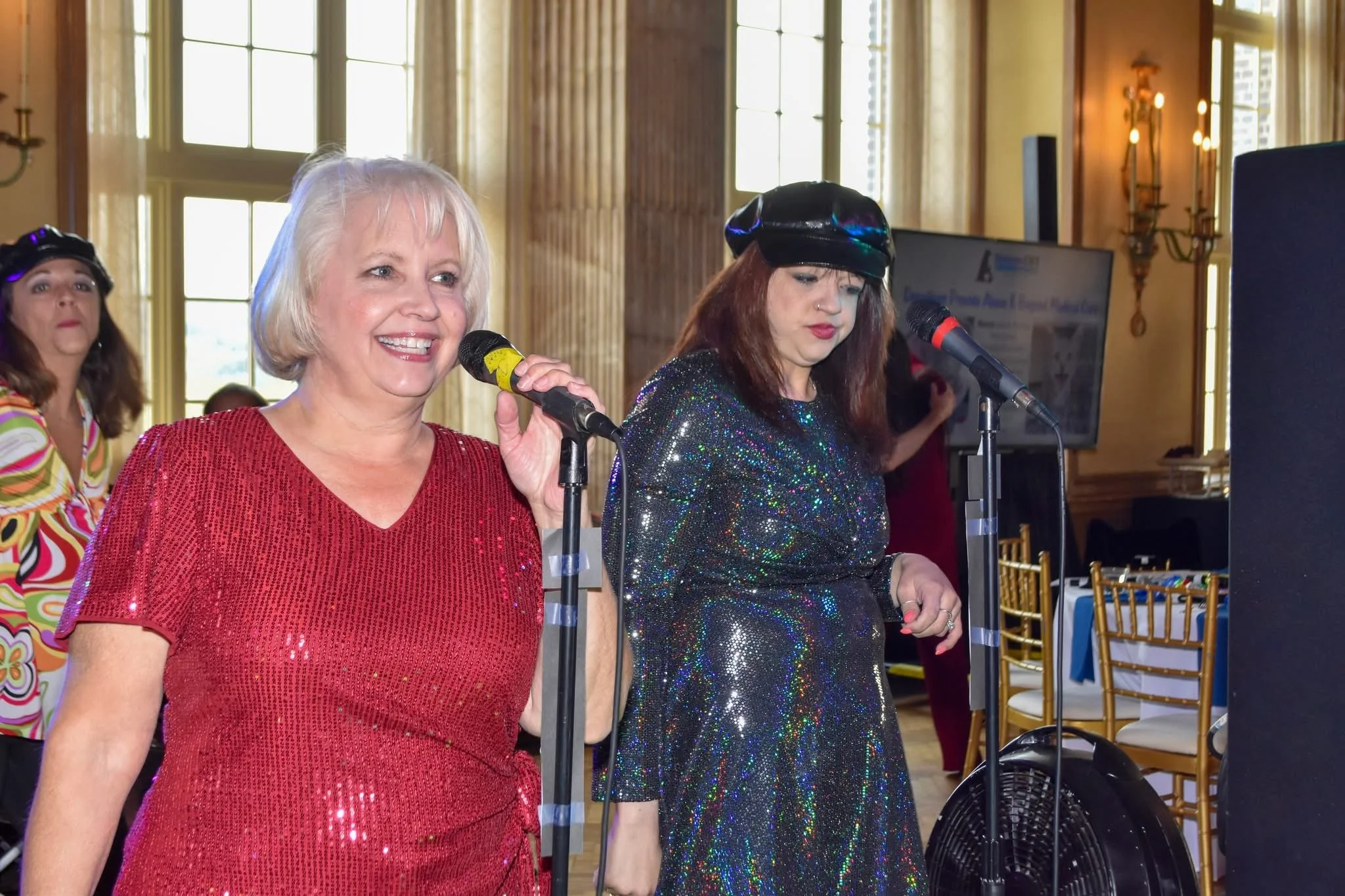 Three women standing at microphones in a decorated indoor setting, dressed in colorful, shiny clothing, with large windows and elegant lighting in the background.