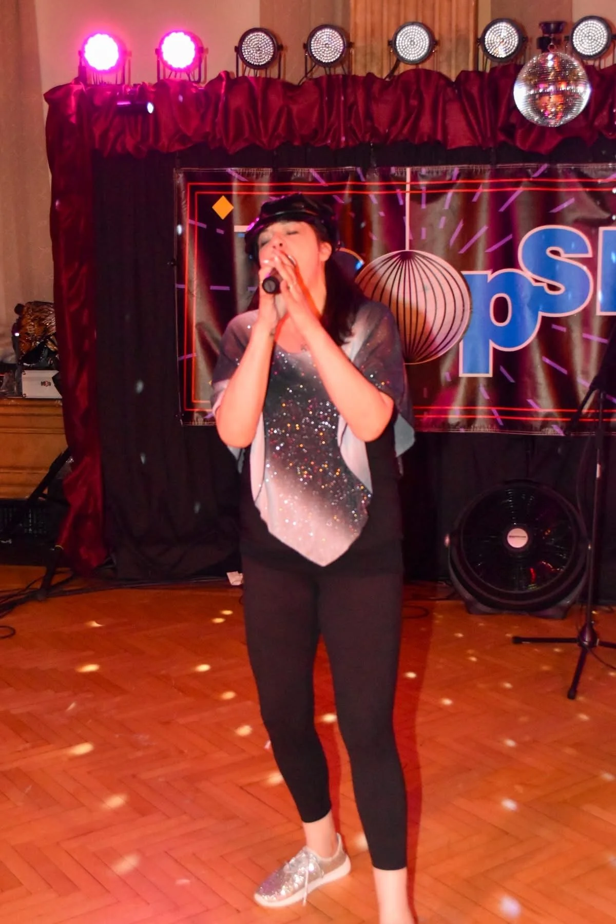 Person singing or speaking into a microphone at a karaoke or stage event, standing in front of a backdrop with colorful lights, a red curtain, and a disco ball, with a wooden floor and small scattered light reflections.