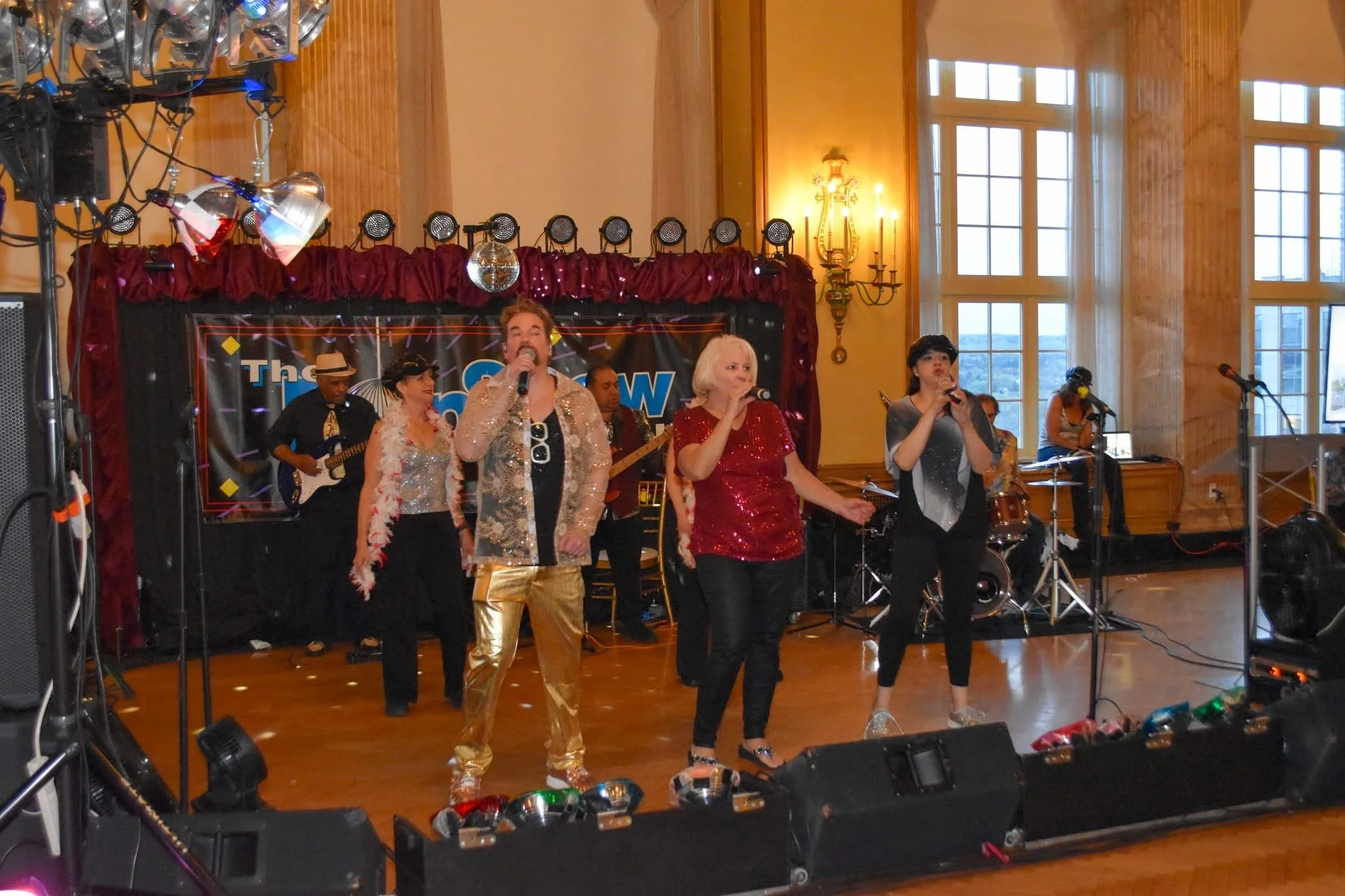 A band of five people performing on stage in a decorated indoor venue, with large windows and wooden walls. The performers are singing into microphones, with musicians playing instruments behind them. The stage has a red curtain background and a disc