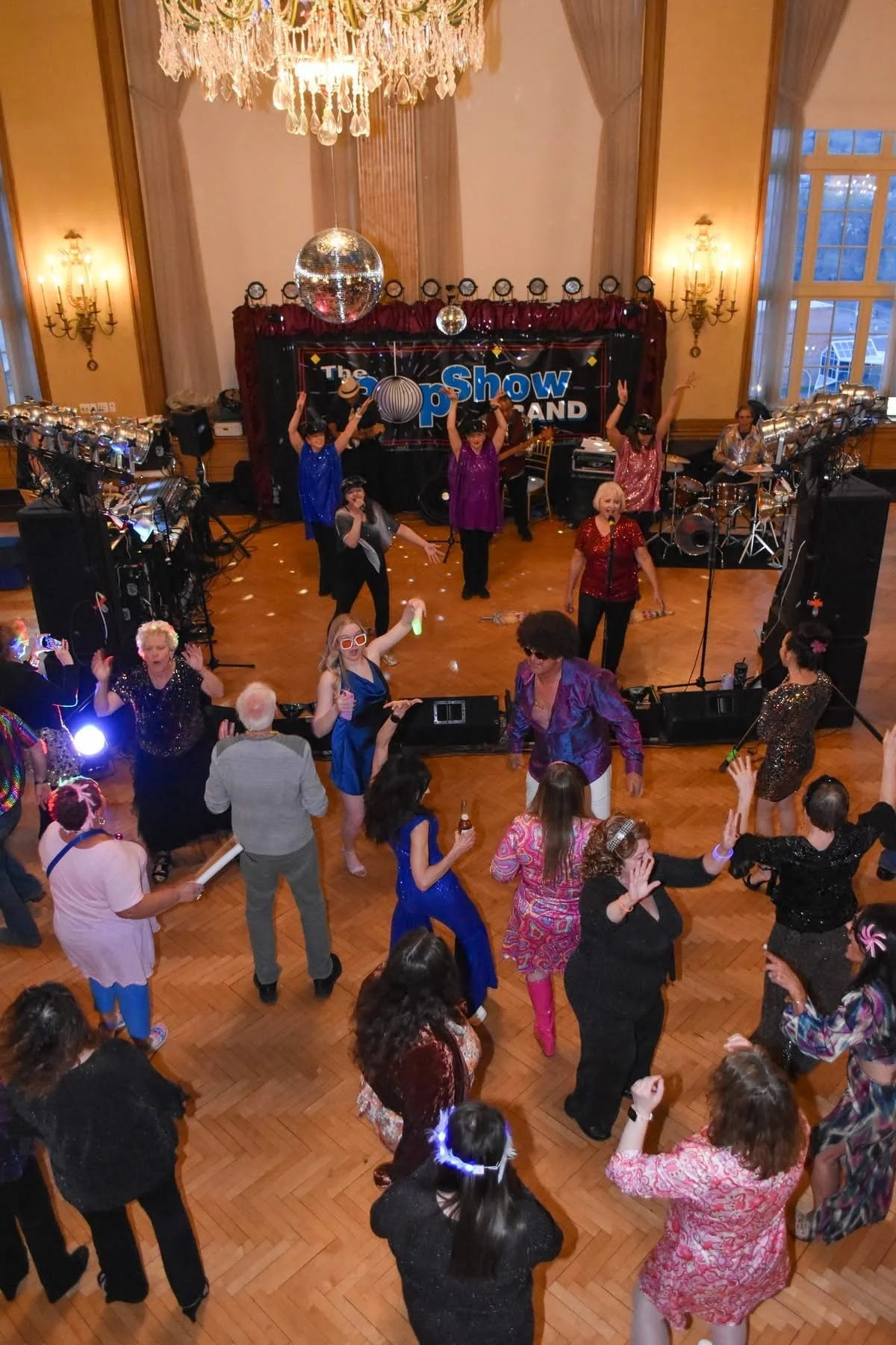 People dancing and enjoying live music at a party with a band performing on stage, disco ball overhead, and the sign 'The Show' in the background.