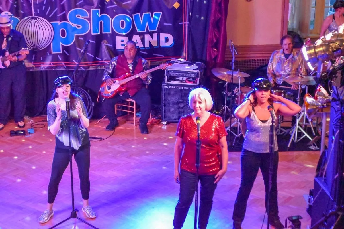 A band performing on stage with three women singing into microphones and three men playing instruments, including guitar, bass, and drums, at a lively indoor concert.