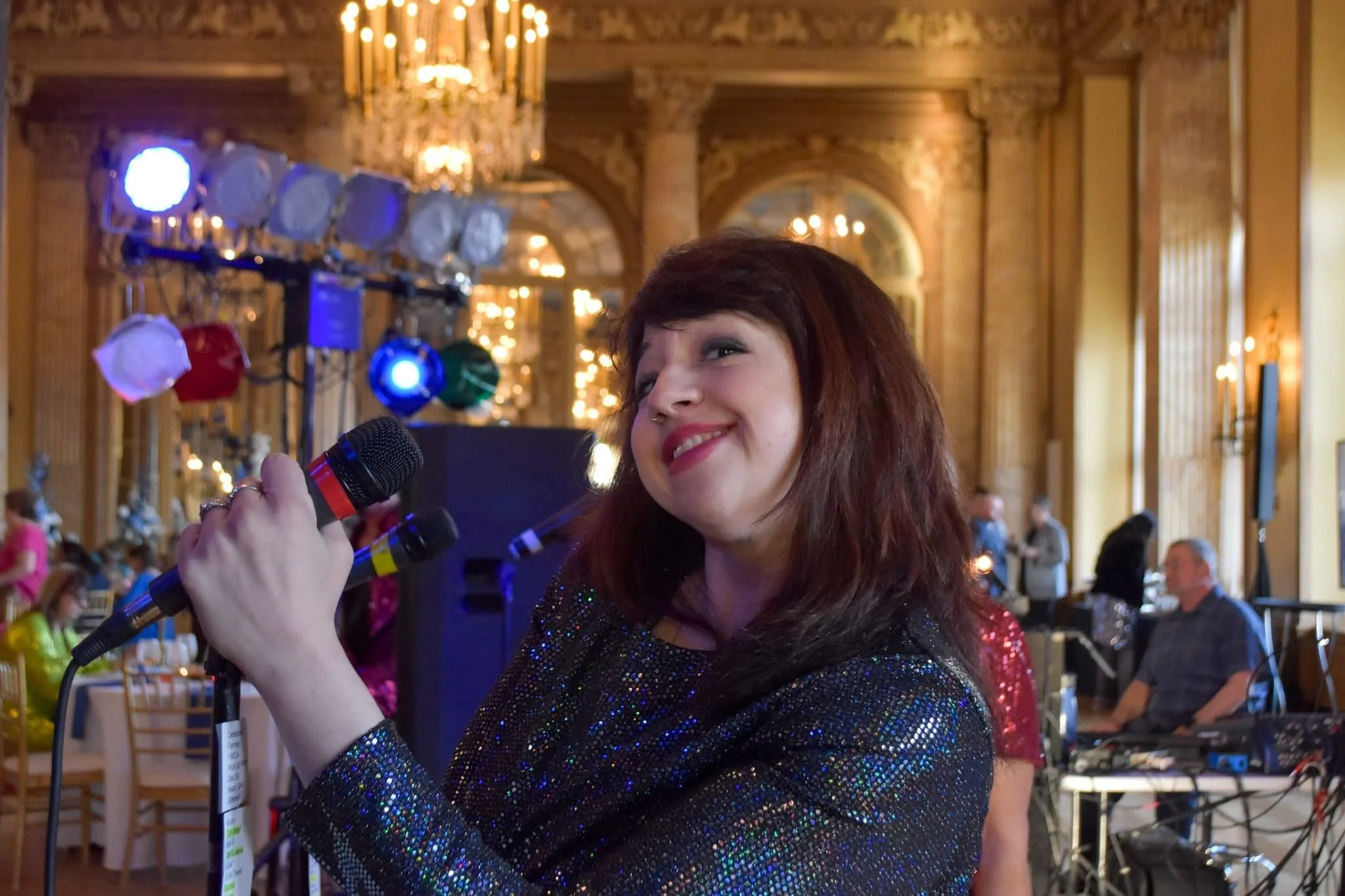 A woman with red hair smiling and holding a microphone at an indoor event with chandeliers, decorative woodwork, and guests seated at tables in the background.