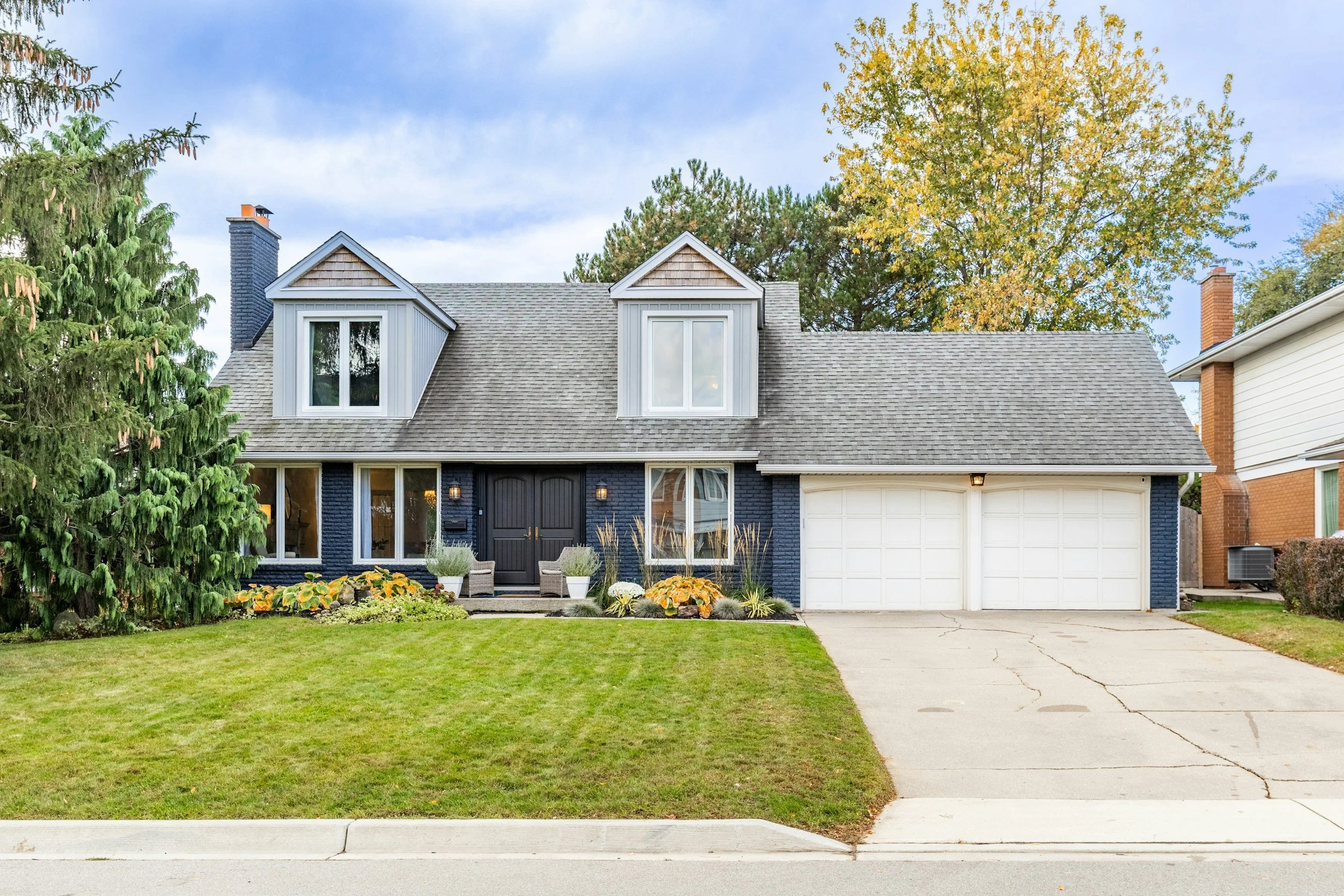 Front view of a two-story house with a grey roof, dark blue brick exterior, white window frames, and a white garage door. The house has a small front porch with two chairs, potted plants, and a lawn with trimmed grass. A large evergreen tree and a deciduous tree with fall foliage are visible in the background.