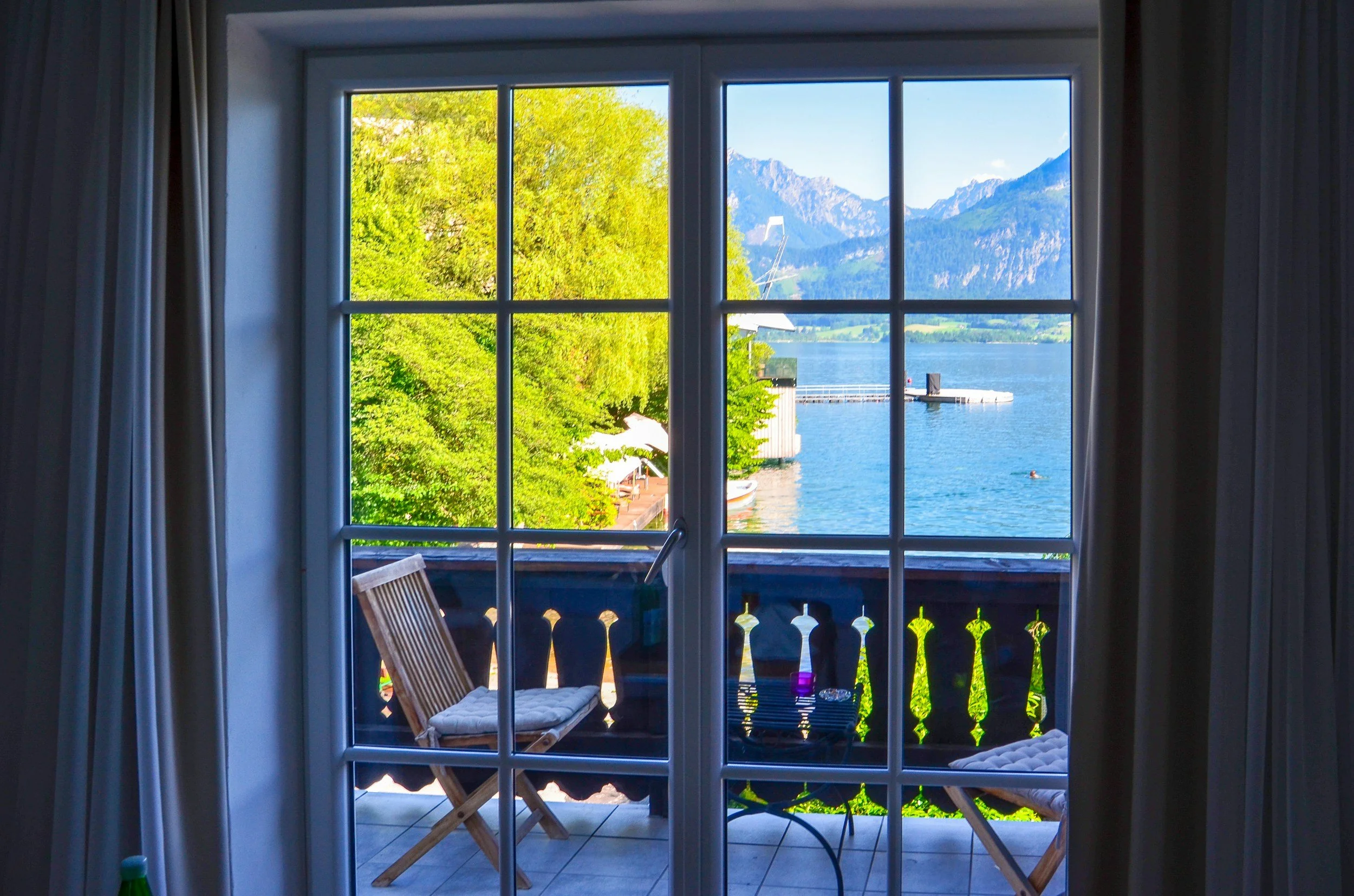 View of a lake and mountains through a glass door with a balcony. The balcony has wooden chairs, a table, and a bottle. Green trees and clear blue sky are visible.
