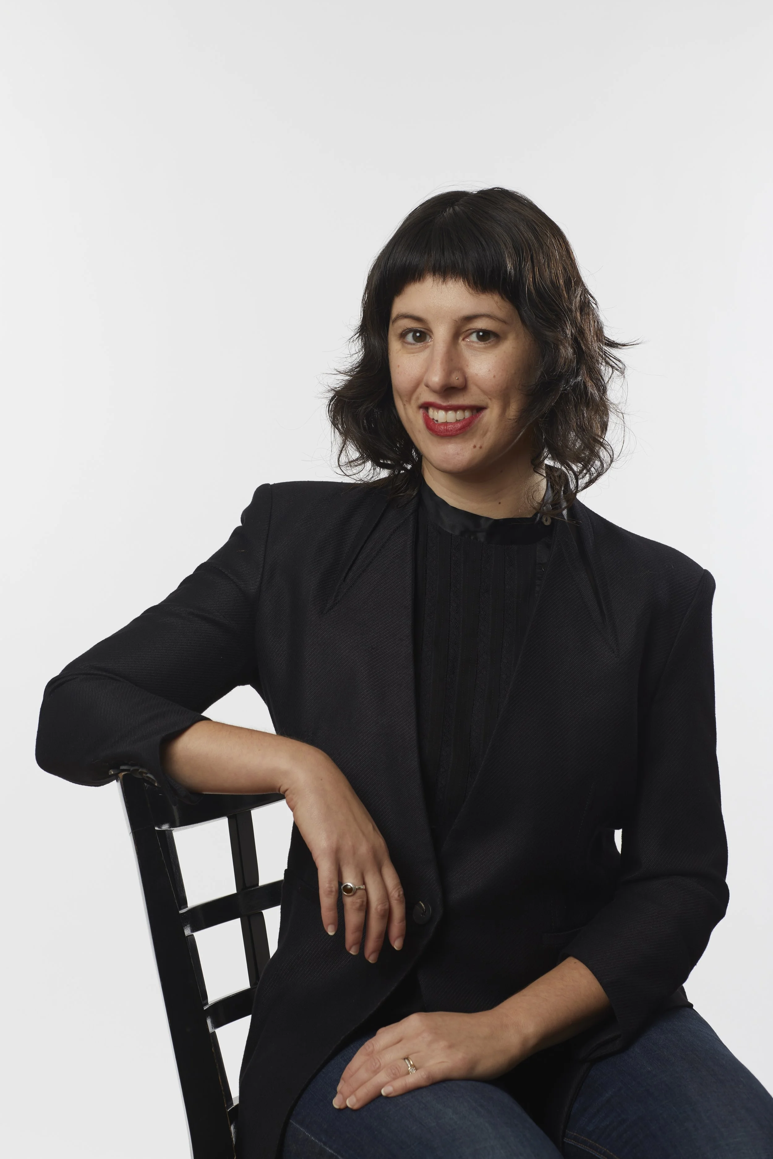 Woman with dark hair and a black blazer sitting on a black chair, smiling, against a plain white background.