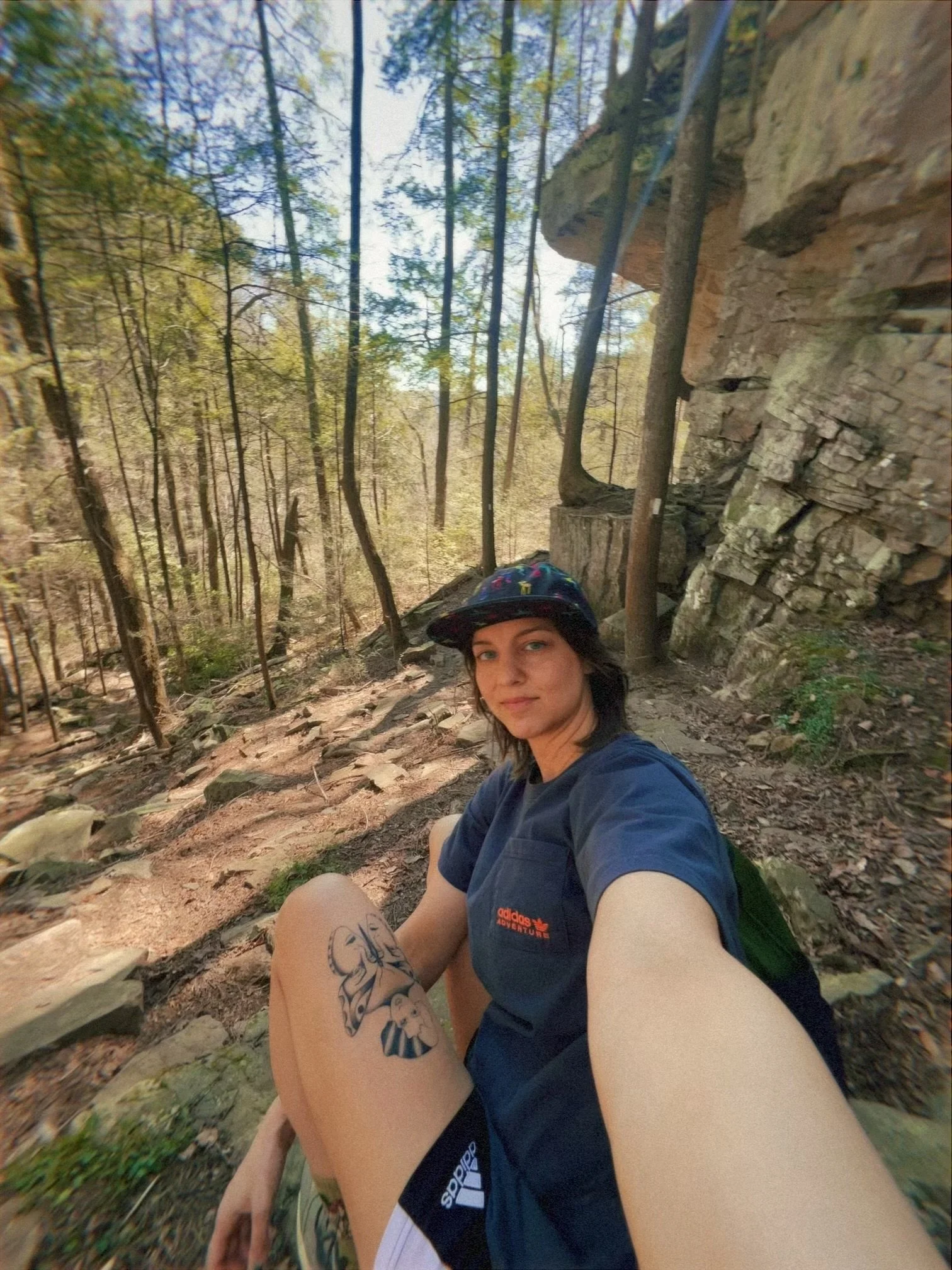 A woman sitting on a dirt trail in a forest, taking a selfie. She has a tattoo on her thigh, wearing a dark hat, a blue t-shirt, and shorts, with trees and rocky terrain around her.