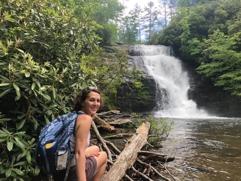 A woman with a backpack sitting on logs by a waterfall in a lush green forest.