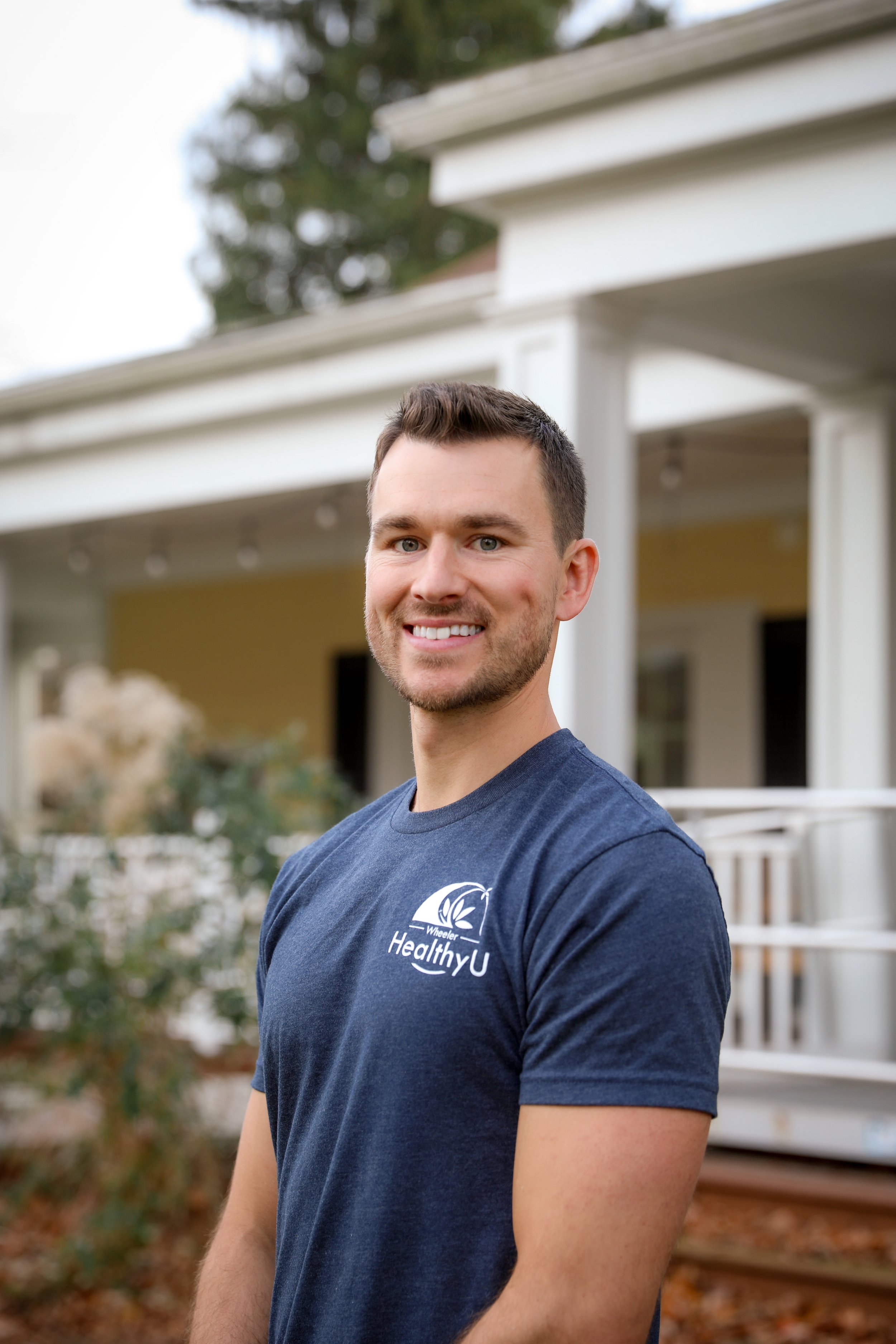 Smiling man wearing a navy blue t-shirt with the 'Wheeler Healthy U' logo, standing outside in front of a house with a porch.