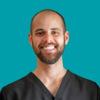 A man with a beard smiling against a teal background, wearing black scrubs.
