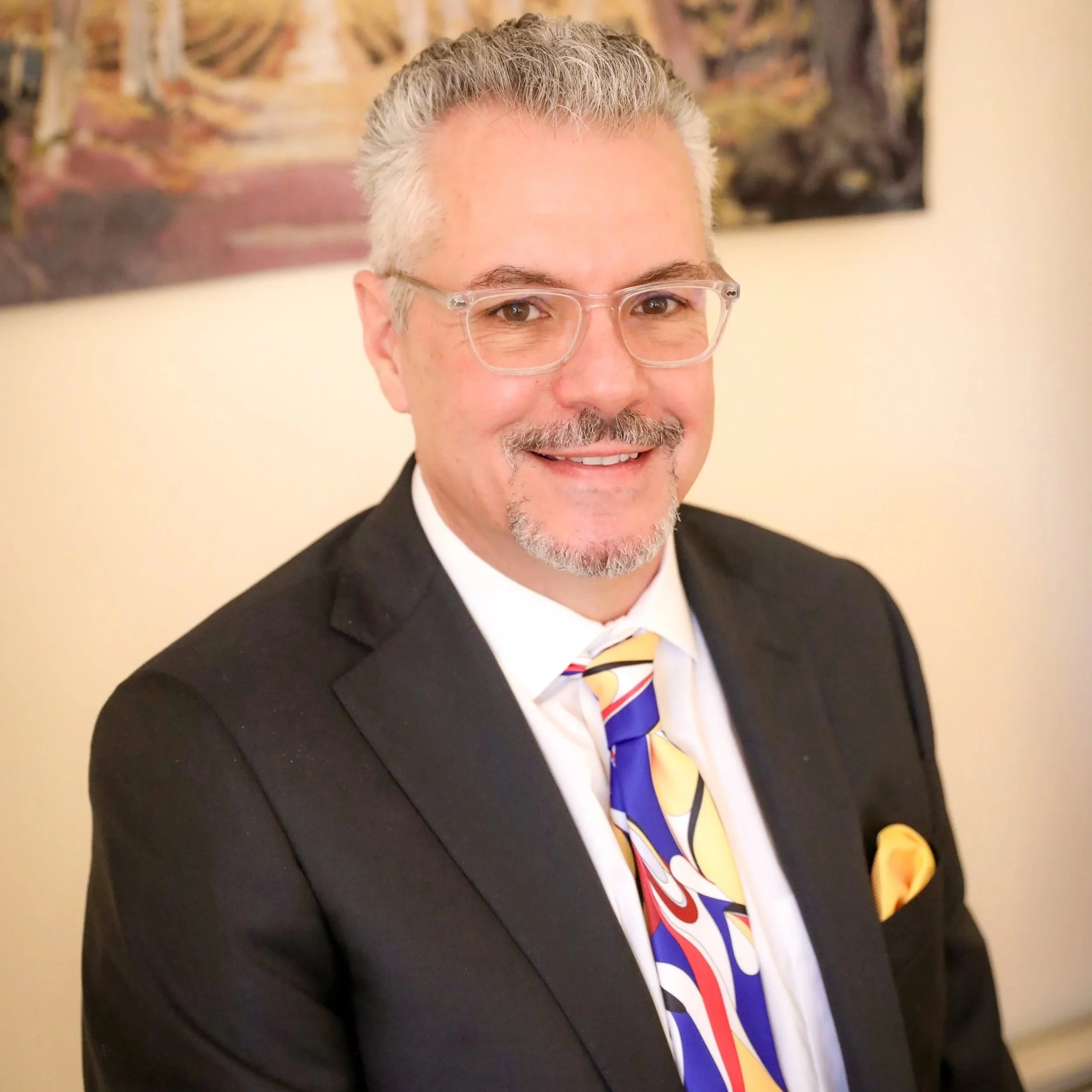 A smiling man with glasses, gray hair, and a goatee, wearing a black suit, white shirt, colorful tie, and a yellow pocket square, standing indoors with a painting in the background.