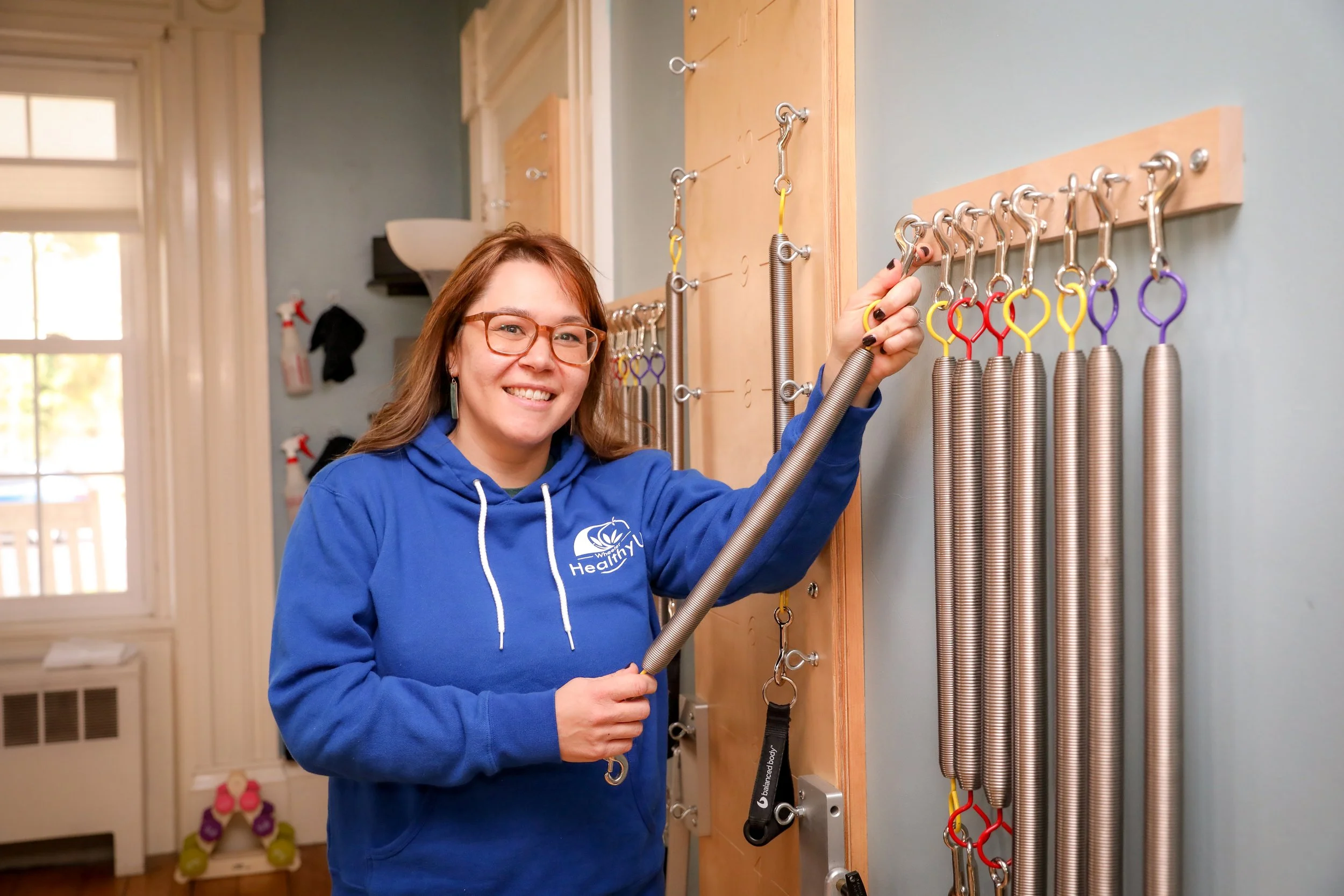 A woman with glasses and a blue hoodie smiling while handling a spring at a therapy or rehabilitation center. She is in front of a wall with several other springs hanging on hooks.