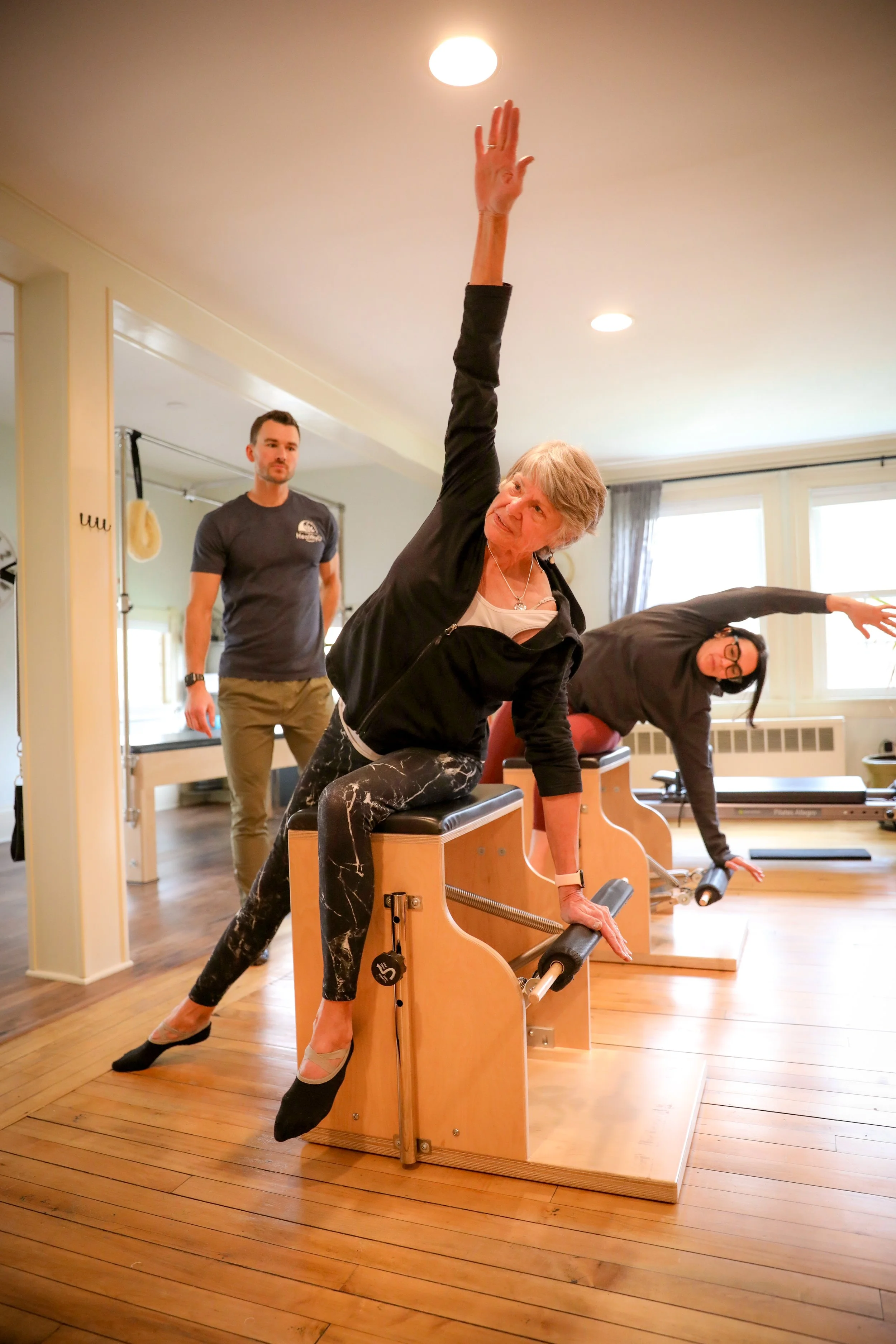 Two women performing Pilates exercises on reformer machines, with an instructor observing in a bright, wooden-floored studio.