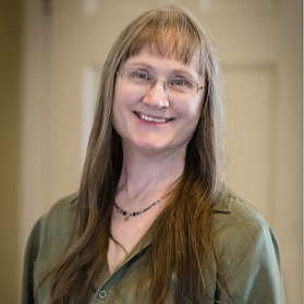 A woman with shoulder-length brown hair and glasses, smiling in an indoor setting.
