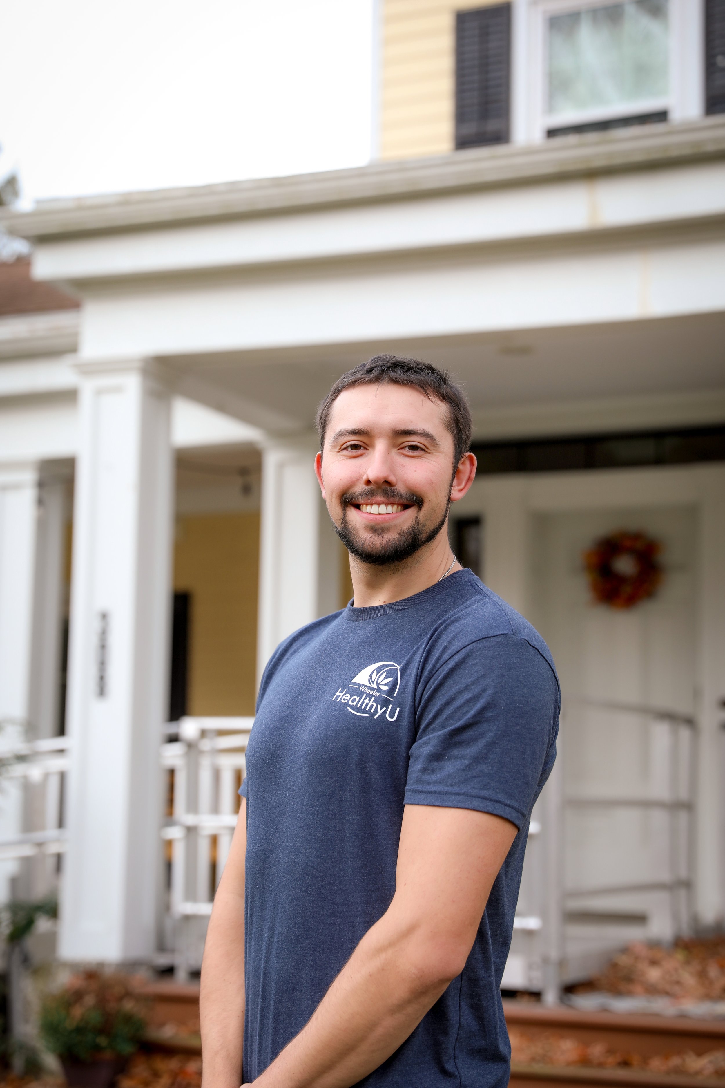 A smiling young man with a beard and short dark hair standing outside a house with a white porch and yellow siding, wearing a navy blue T-shirt with "Healthy U" text and logo.