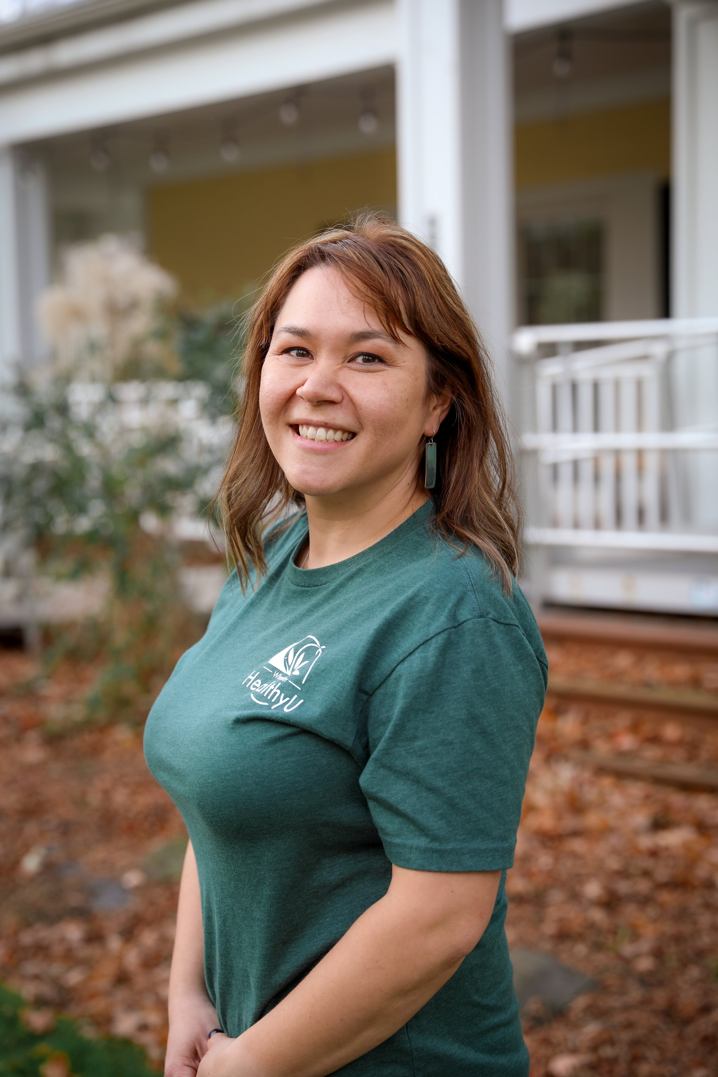 A woman with shoulder-length brown hair smiling outdoors, wearing a green T-shirt with a 'HealthyU' logo, standing in a yard with autumn leaves and a house with a porch in the background.