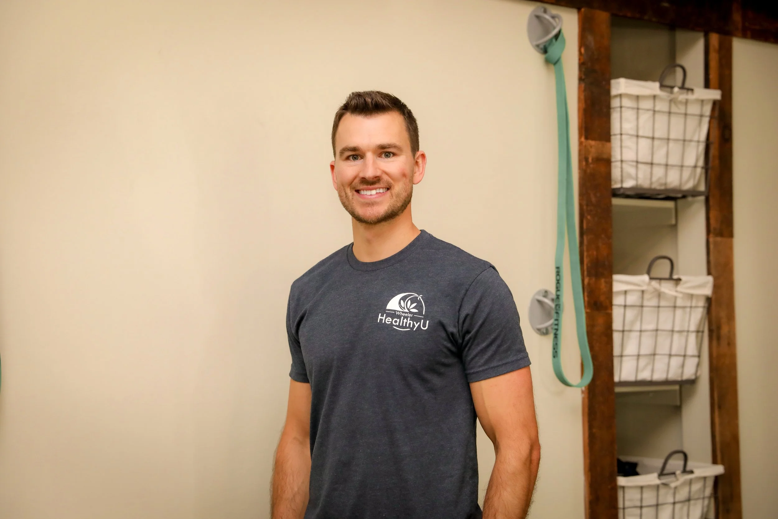 A smiling man with short dark hair and a beard wearing a dark gray t-shirt with 'HealthyU' logo, standing indoors against a light-colored wall with shelves in the background.