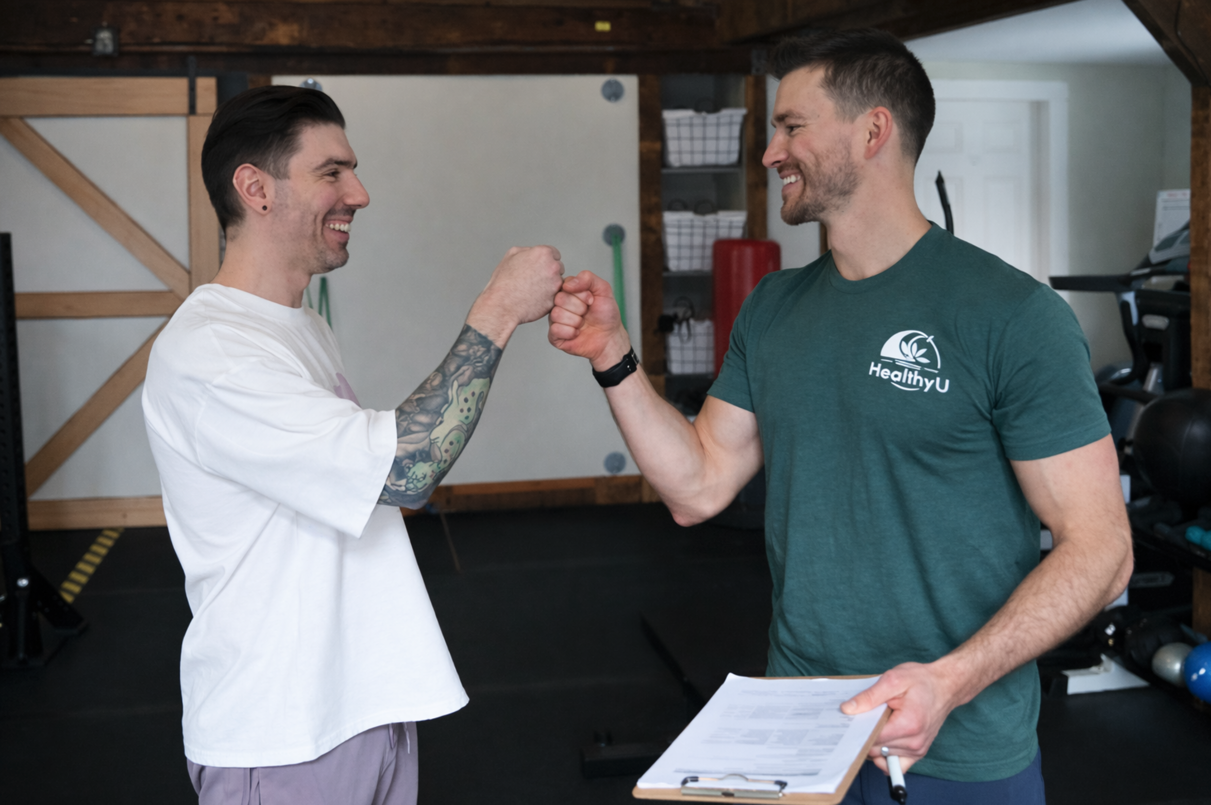Two men are fist-bumping in a gym, smiling at each other. One man has tattoos on his arm and is wearing a white t-shirt, while the other is wearing a green shirt with a logo and holding a clipboard.