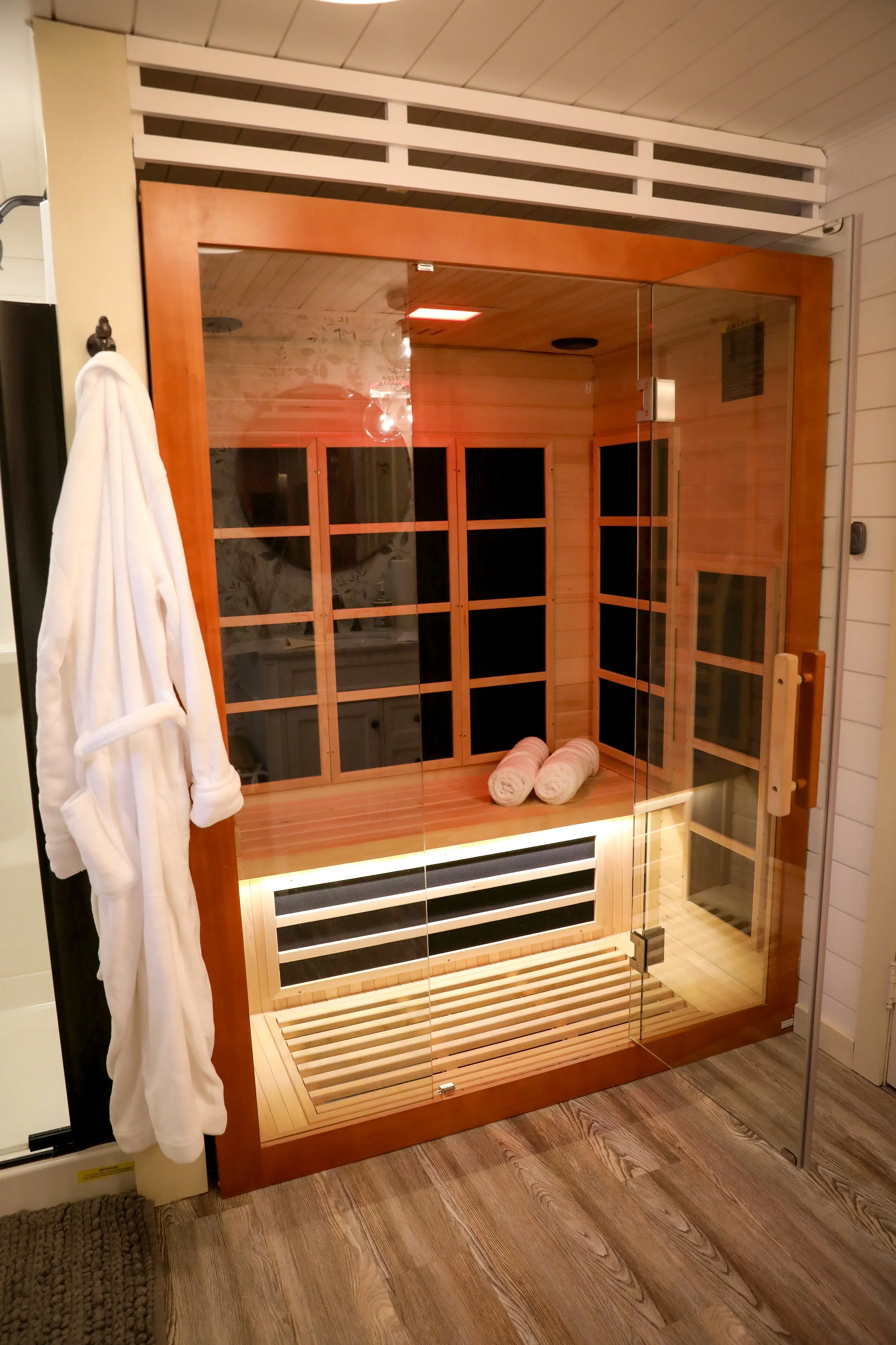 A glass-enclosed infrared sauna with wooden interior, two rolled towels inside, a white bathrobe hanging outside, wood flooring, and a vent or heater at the bottom.