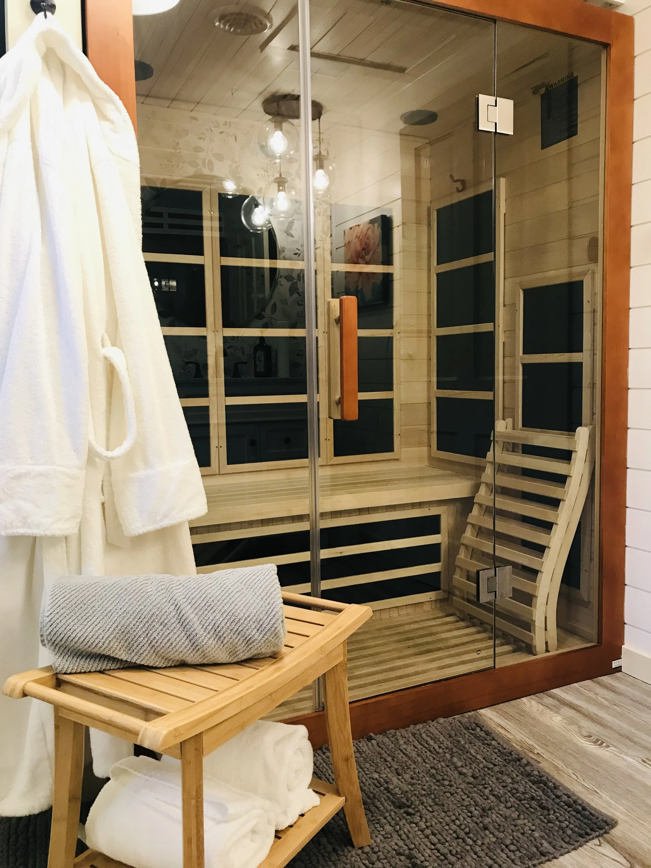 A glass-enclosed sauna room with a wooden bench and a wooden folding chair inside. Outside the sauna, a white bathrobe hangs, and on a small wooden stool, there is a rolled-up towel. The room has wood paneling and a textured rug on the floor.
