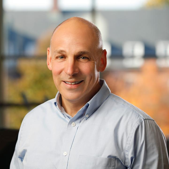 A smiling middle-aged man with a bald head wearing a light blue dress shirt in an indoor setting with blurred windows and trees in the background.