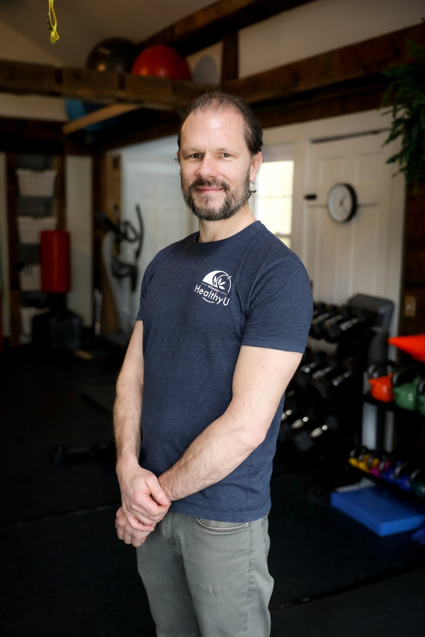 A man standing in a gym, wearing a navy Blue T-shirt with the words 'Healthy U' and a logo, with gym equipment visible in the background.