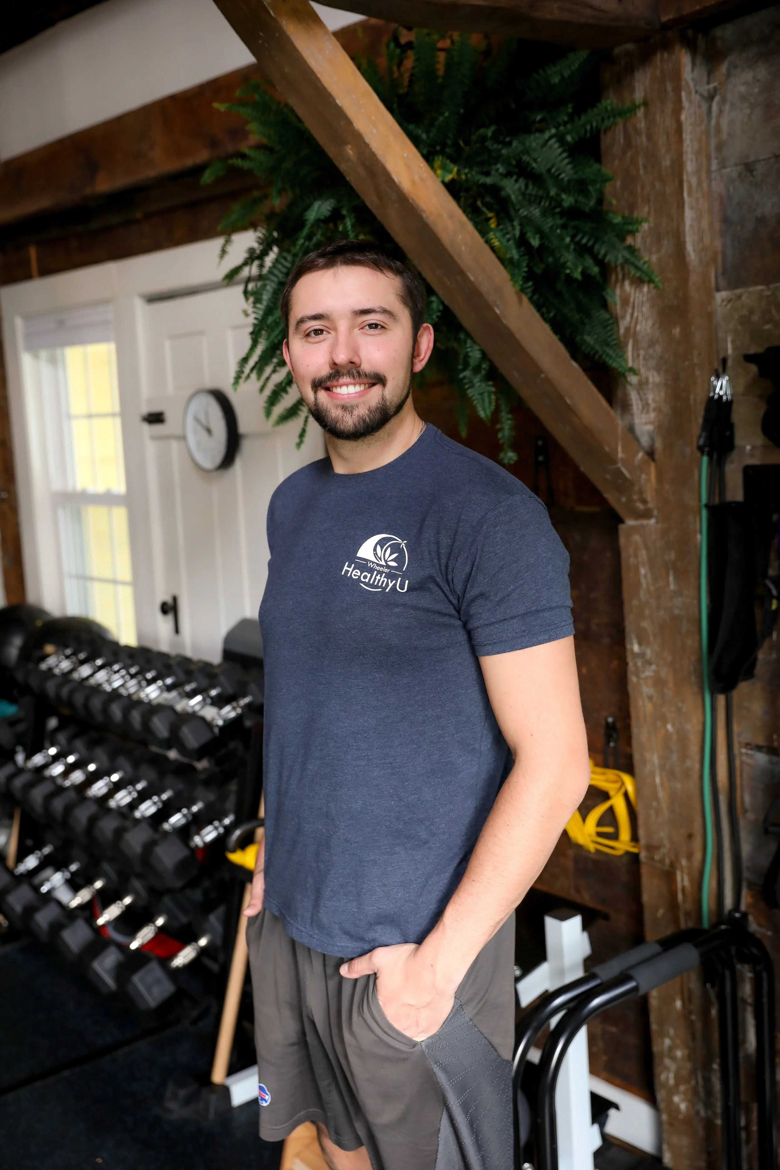 A smiling man with a beard and short dark hair standing indoors, wearing a navy t-shirt with a logo that says 'Healthy U.' Behind him, there are two windows, a wall clock, a set of dumbbells, and various workout equipment in a rustic room with wood-paneled walls and a large plant.