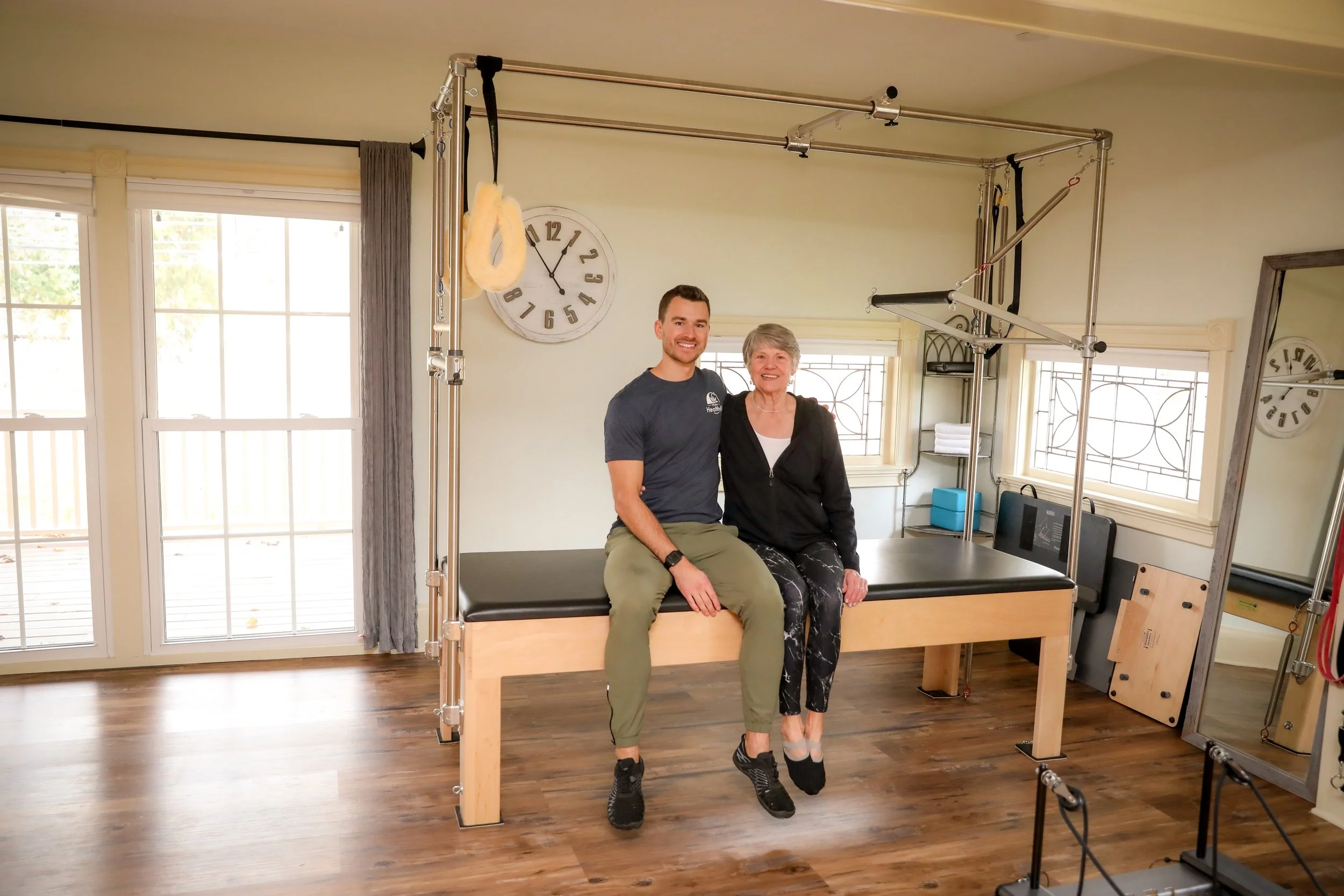 A young man and an older woman sitting on a Pilates reformer in a bright exercise room with large windows, wooden floor, and a wall clock.