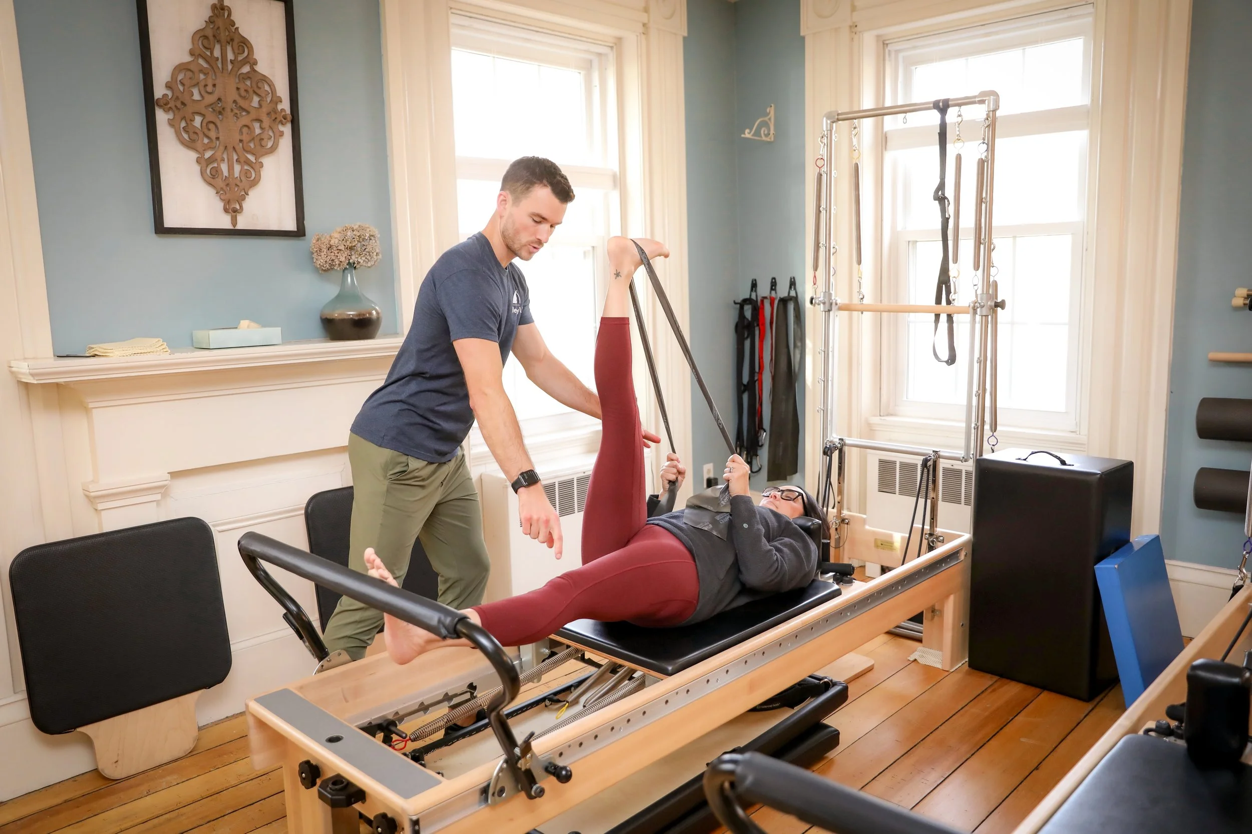 A woman is lying on a Pilates reformer machine with her legs raised at a 90-degree angle, holding straps with her hands, while a trainer assists her by holding her leg. The room has wooden floors, large windows, and Pilates equipment.