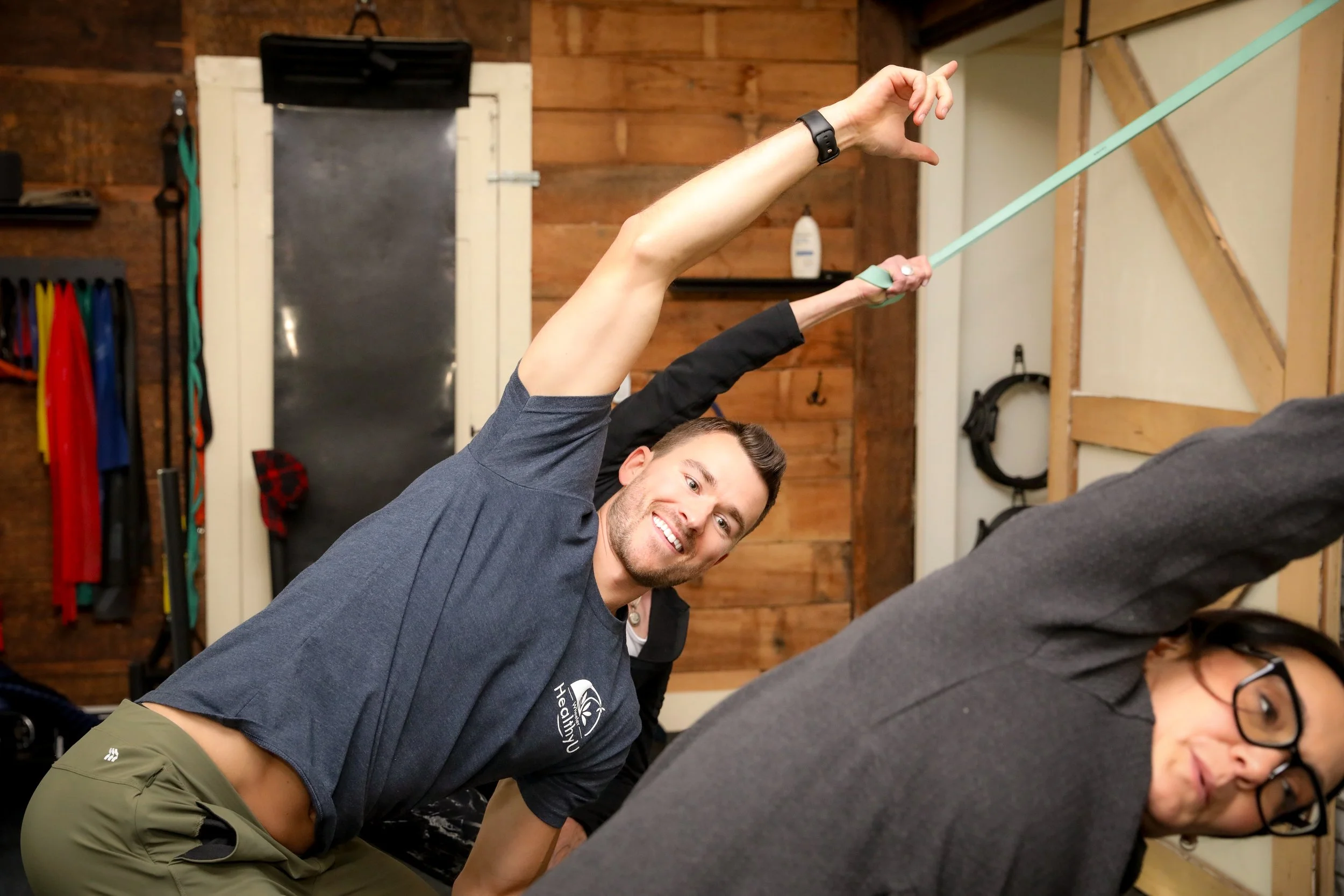 A man smiling and stretching with a resistance band in a rustic gym or workout space, with a woman in front doing a stretch, both engaging in fitness activities.