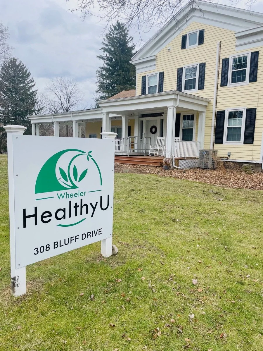 Sign for Wheeler HealthyU at 308 Bluff Drive in front of a yellow house with a porch and black shutters, surrounded by grass and leafless trees.