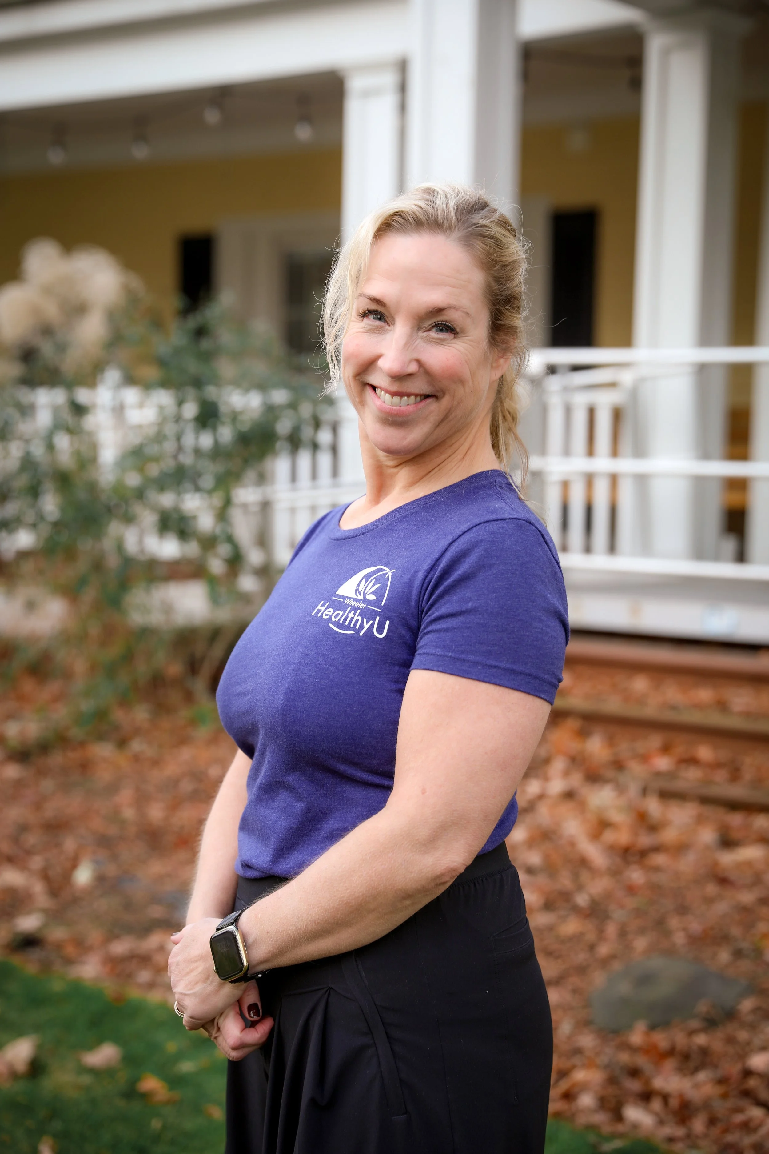 Smiling woman wearing a blue T-shirt with the 'HealthyU' logo, standing outside near a house with a porch and autumn-colored leaves on the ground.