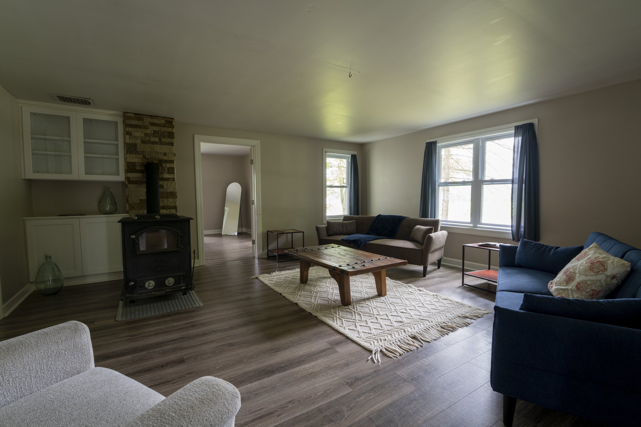 Living room with hardwood floors, three windows with blue curtains, a wood coffee table, a beige sofa, a blue armchair, and a white armchair, with a wood stove and brick chimney on the left