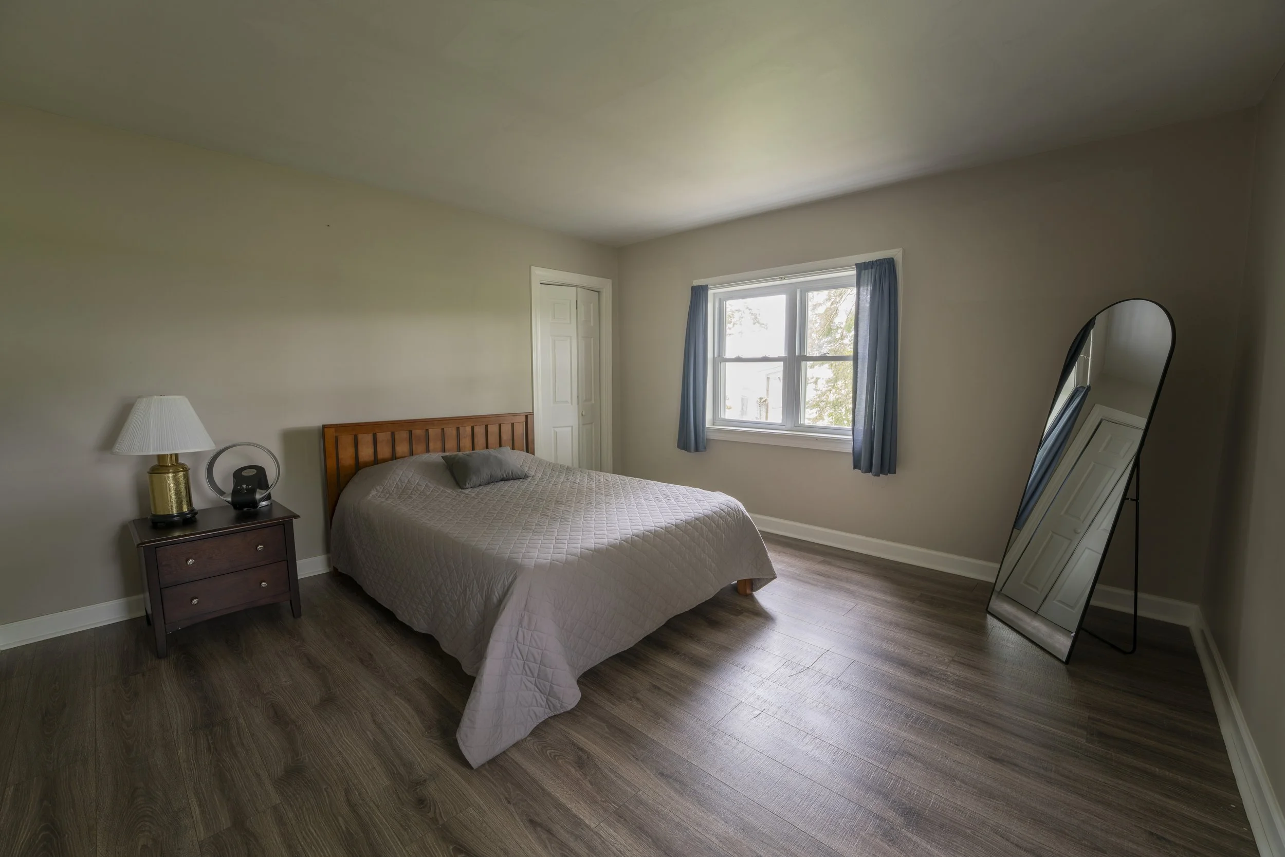 A bedroom with a bed, nightstand, lamp, mirror, and window with blue curtains.