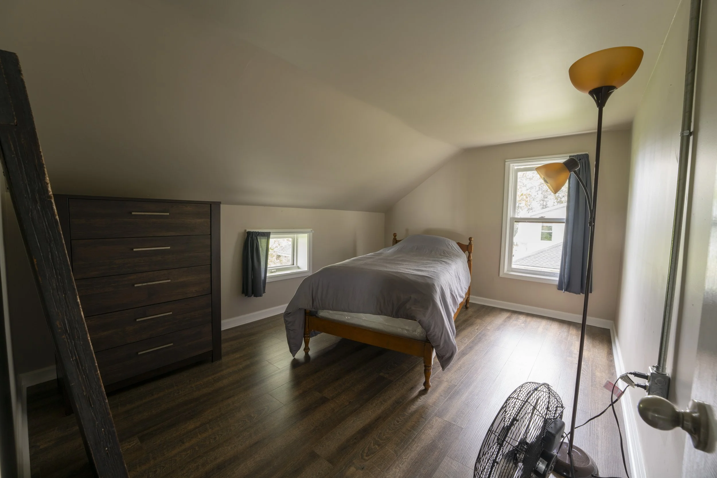 A small bedroom with a sloped ceiling, painted walls, two windows with blue curtains, a wooden bed with a gray duvet, a dark wooden dresser, a standing lamp with two yellow lampshades, a fan, and hardwood flooring.