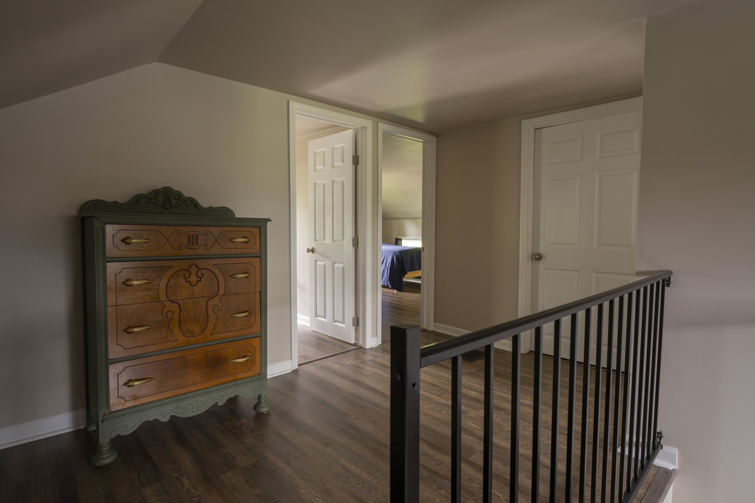 Interior view of a upstairs landing with hardwood floors, a vintage dresser on the left, white doors leading to bedrooms, and a black metal railing.