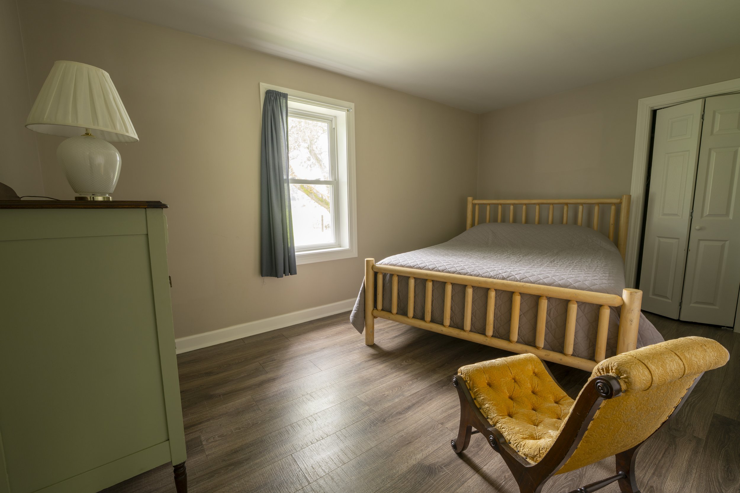 A bedroom with a window, a wooden bed with a gray quilt, a yellow upholstered vintage armchair, a pale green dresser with a white lamp, and beige walls.