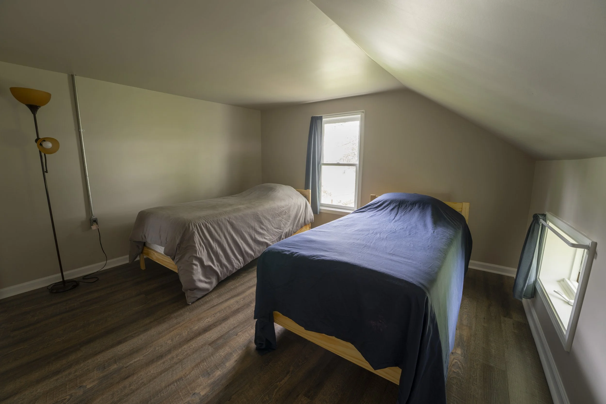 A bedroom with two single beds under a sloped ceiling, one with a beige bedspread and the other with a dark blue cover, window with a blue curtain between the beds, and a floor lamp in the corner.
