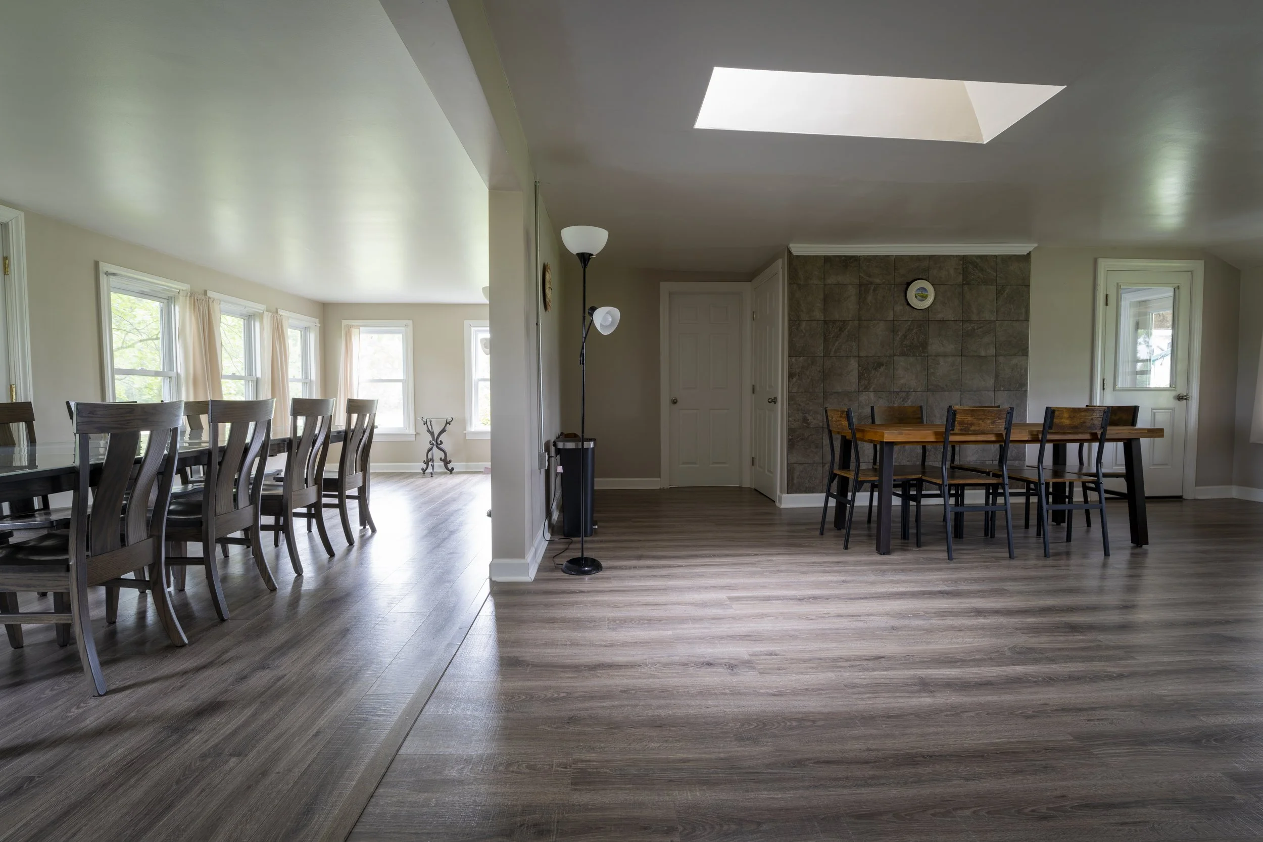 Interior of a house showing a dining area with a large wooden dining table and eight chairs, and a bright, sunlit living area with a row of windows, beige curtains, and hardwood floors.