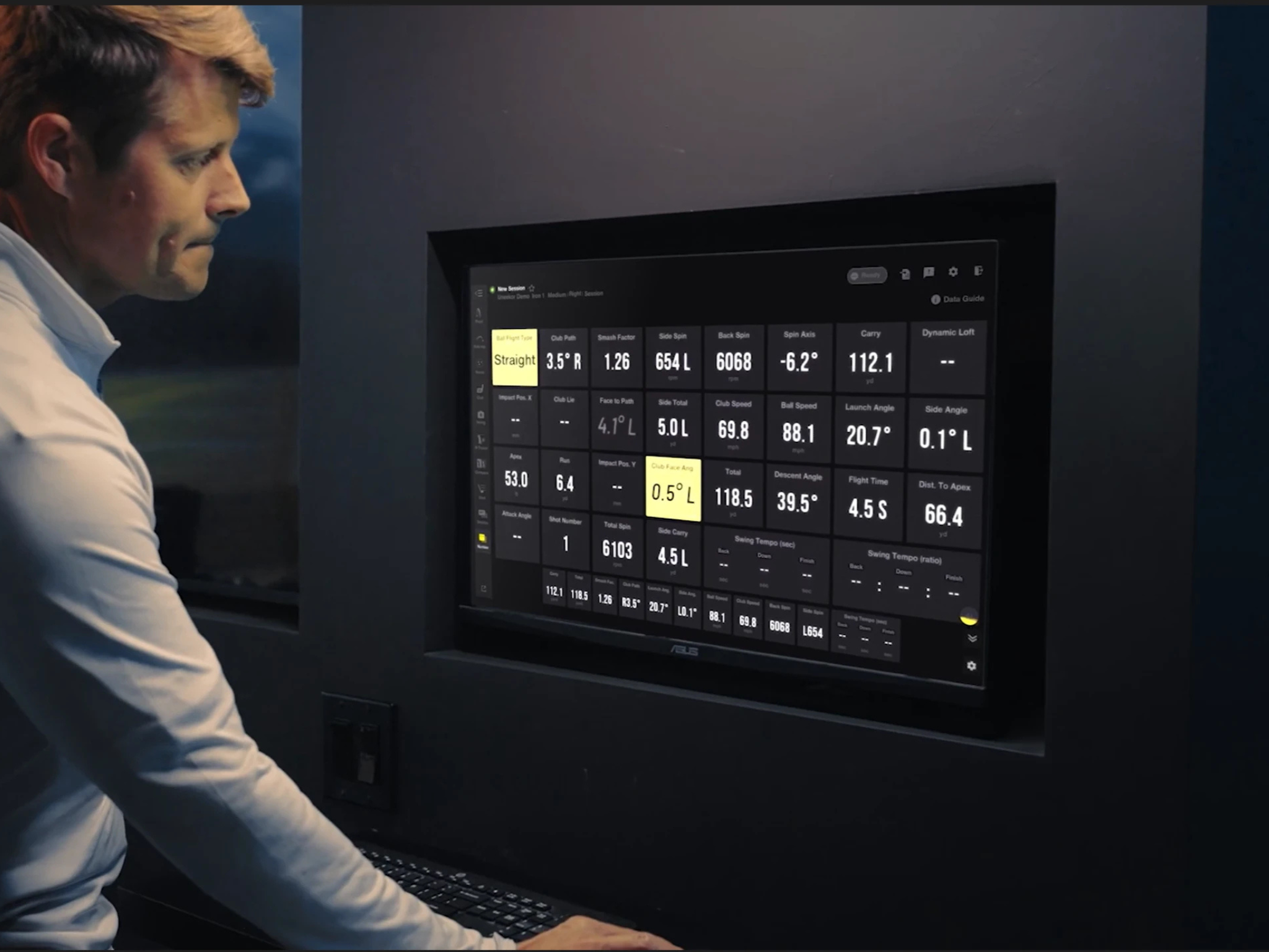 Man in a white shirt looking at a golf simulation data screen showing various golf metrics.