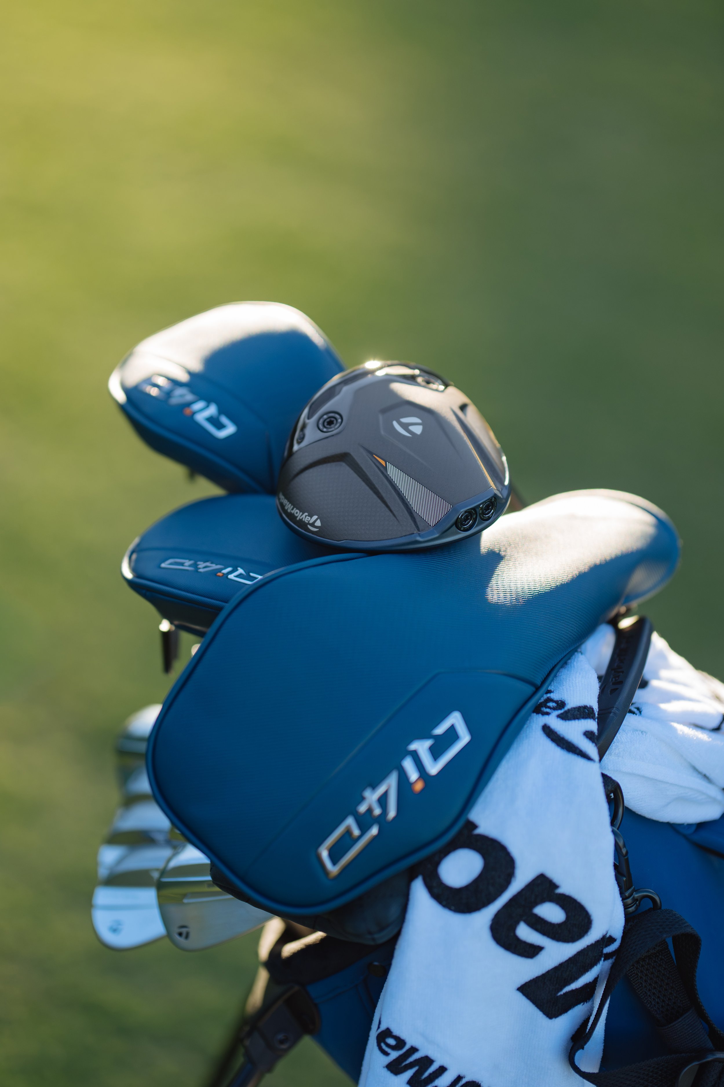 Golf driver club head resting on a golf bag with golf clubs and a towel on a golf course.