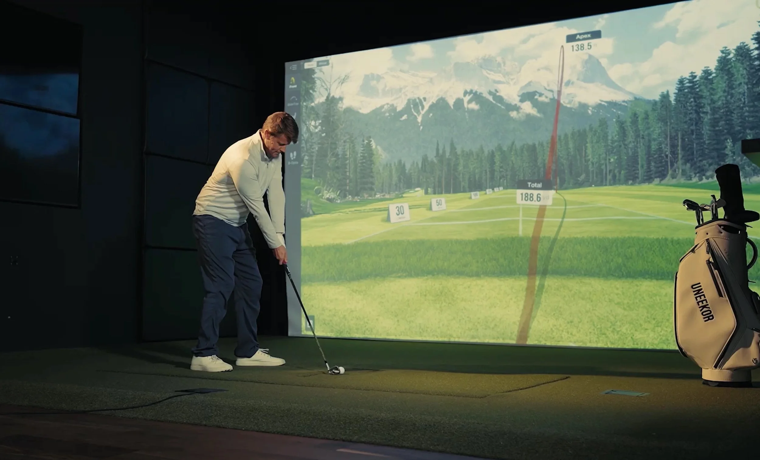 Man practicing golf indoors at a simulator with a large screen displaying a mountain landscape and a golf course.