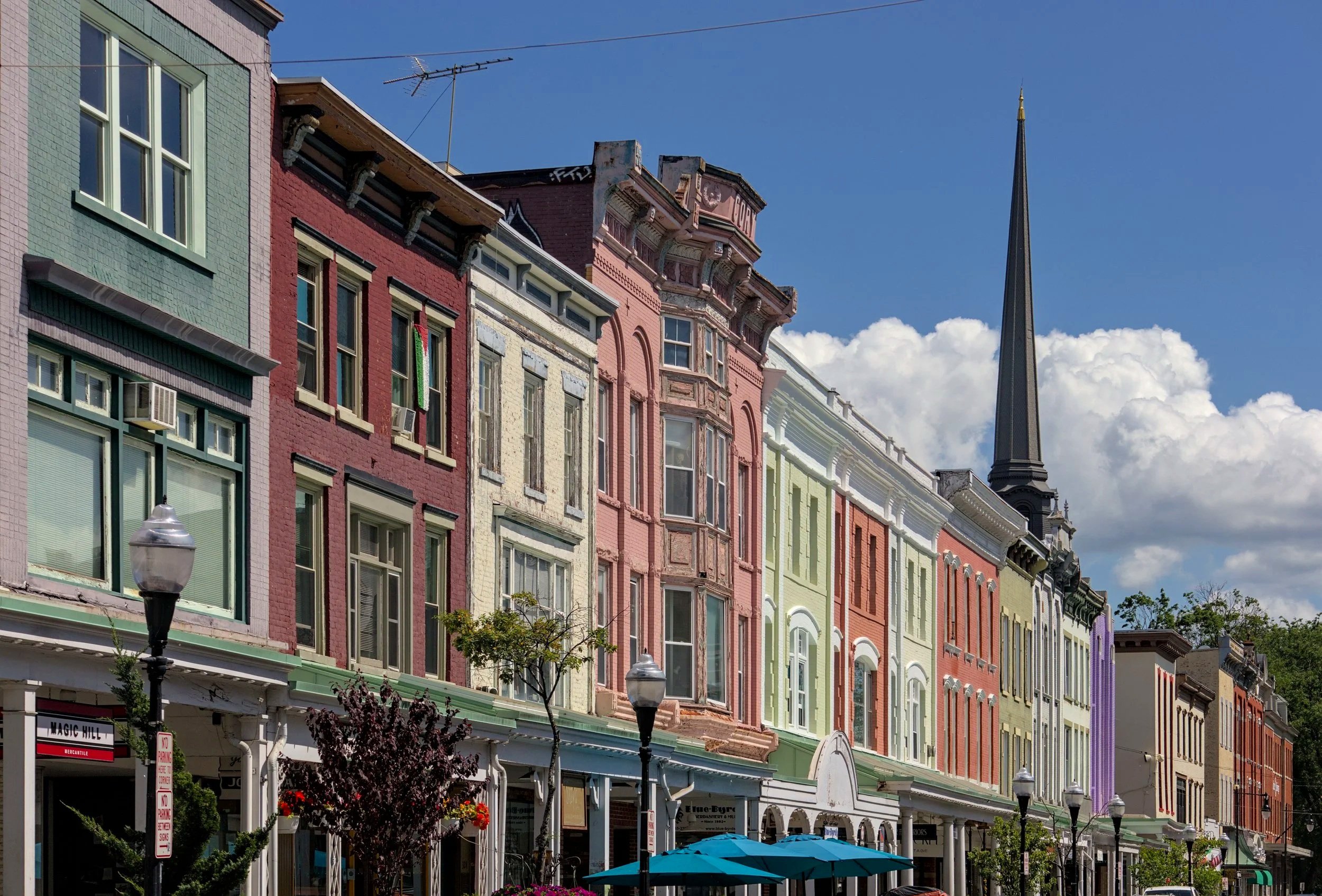 Colorful Victorian-style commercial buildings along a street with trees, street lamps, and storefronts under a partly cloudy sky.