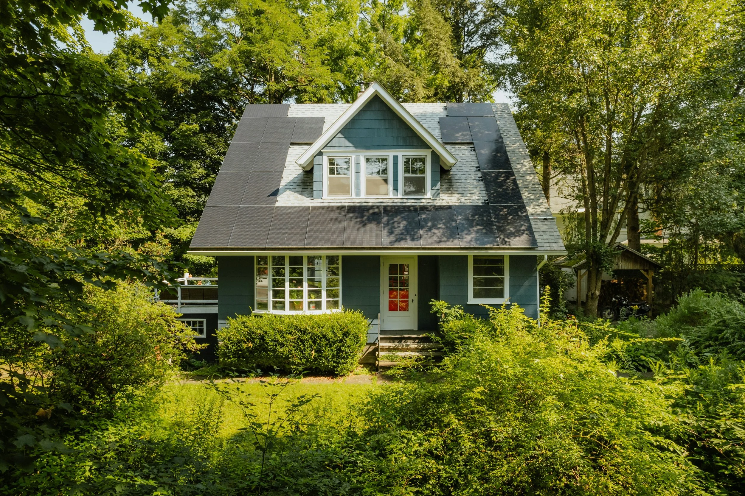 A blue house with a gable roof and large front windows, surrounded by green bushes and trees, with solar panels on the roof.