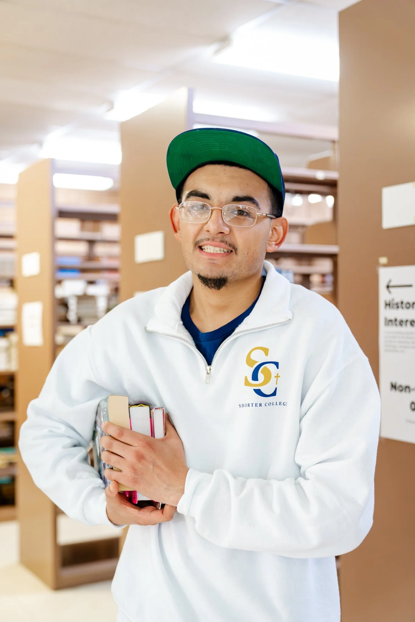 A college student holding books in a library, wearing glasses, a white hoodie with a college logo, and a green cap.
