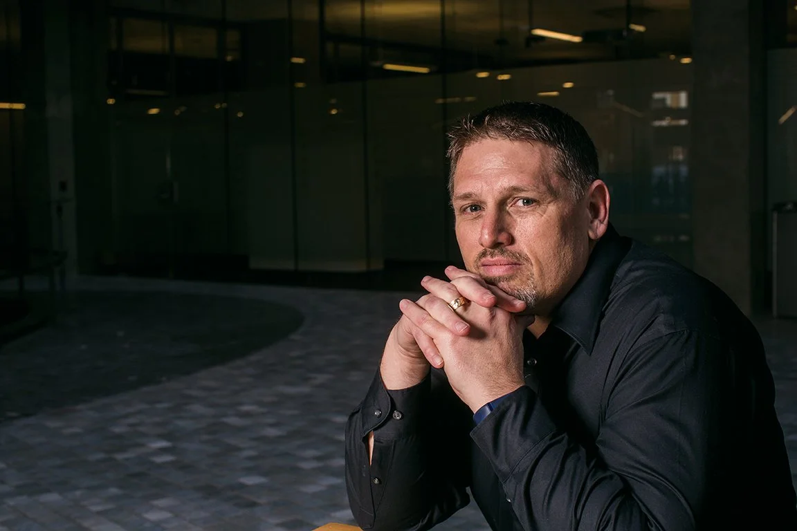 A man with short dark hair, a goatee, and wearing a black shirt, smiling with arms crossed, leaning against a concrete wall.