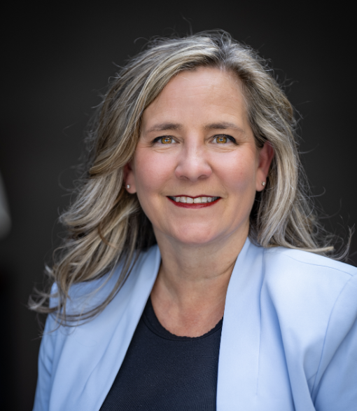 A woman with long brown hair smiling outdoors, wearing a black shirt and a silver pendant necklace, with green foliage and trees in the background.