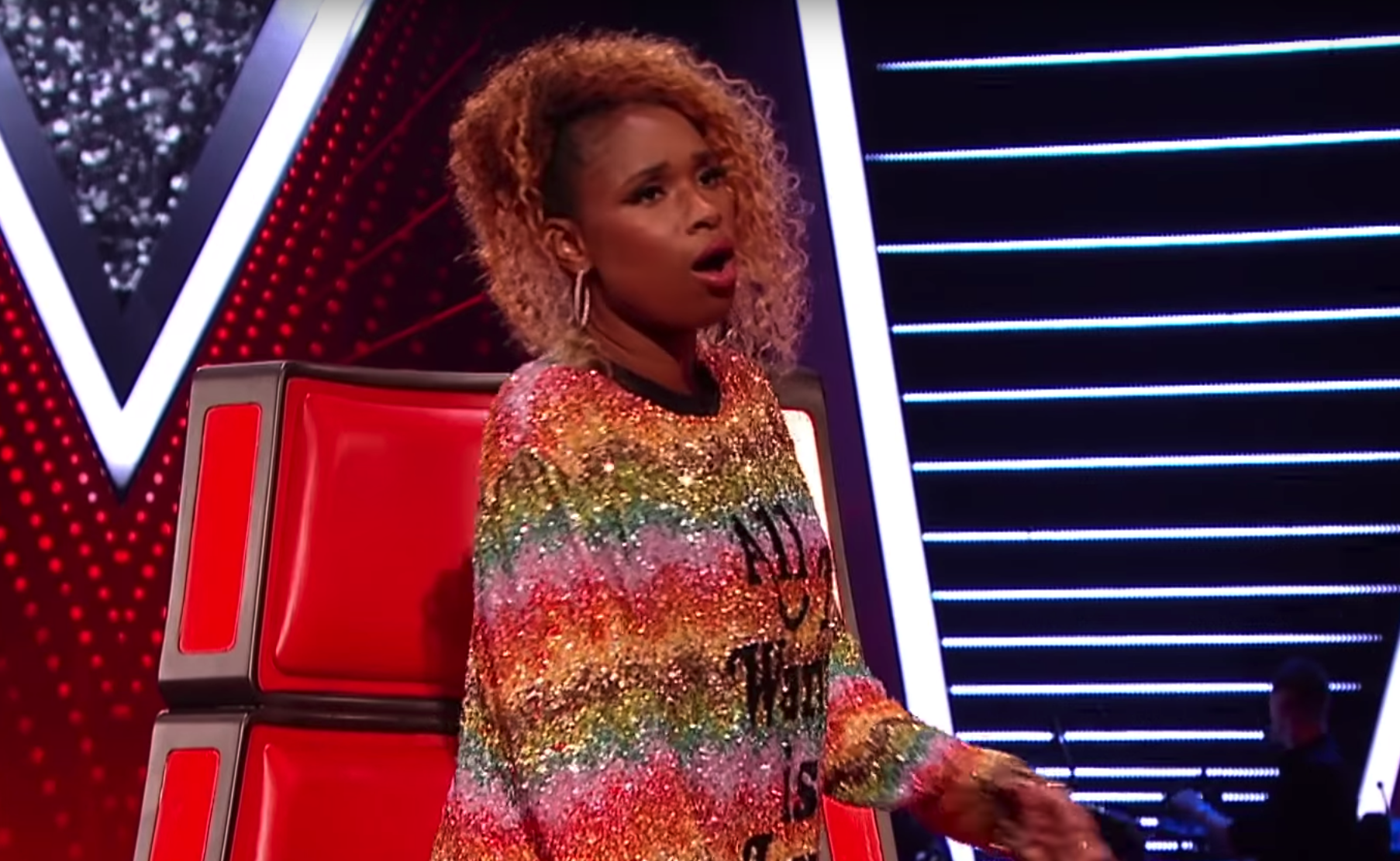 A woman with curly, blonde hair sitting in a red chair on a stage with a brightly lit, modern background.