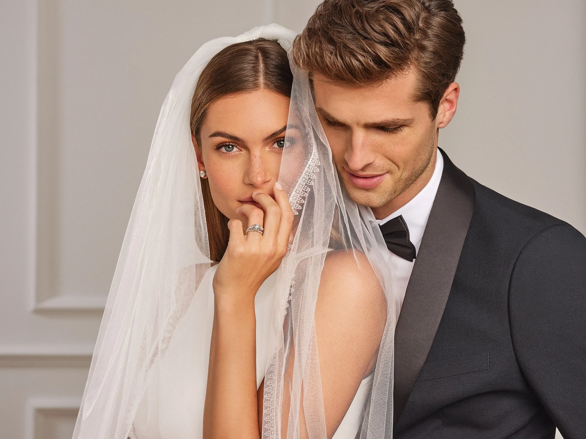 A bride and groom pose together, with the bride partially covering her face with her veil, and the groom looking down with a slight smile.