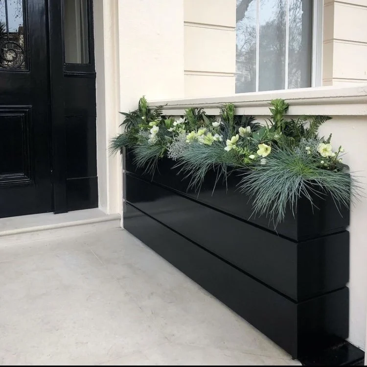 A porch with a hanging decorative rug featuring geometric patterns, overlooking a snowy yard with a deer resting near a big evergreen tree, and a house with a wood-paneled ceiling.