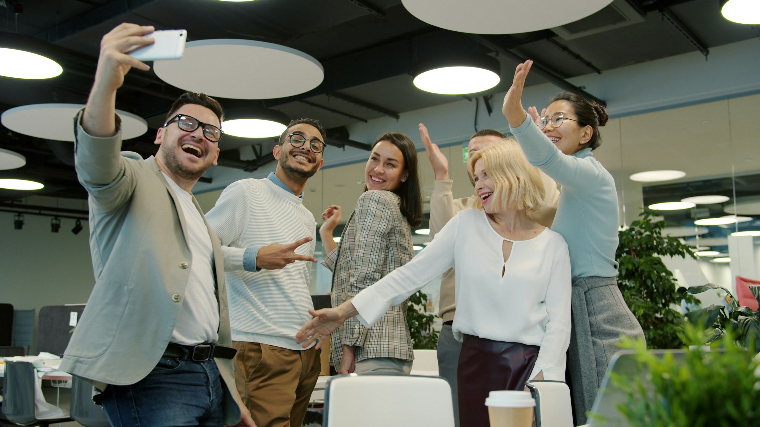 Group of five diverse coworkers in casual business attire taking a selfie and smiling in a modern office.